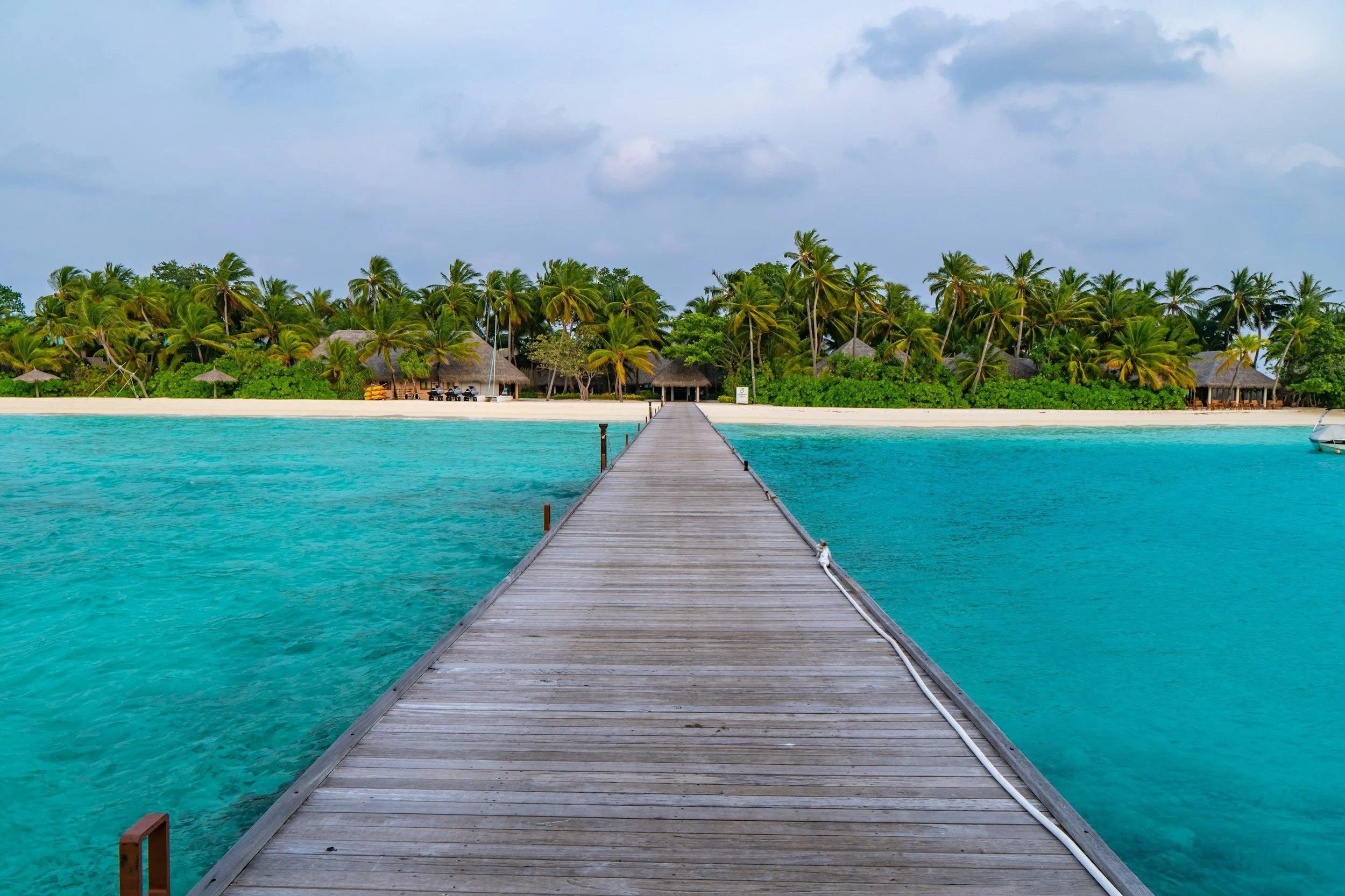 A wooden pier extending over clear turquoise water towards a tropical island with lush green palm trees, beachside huts, and a cloudy sky.