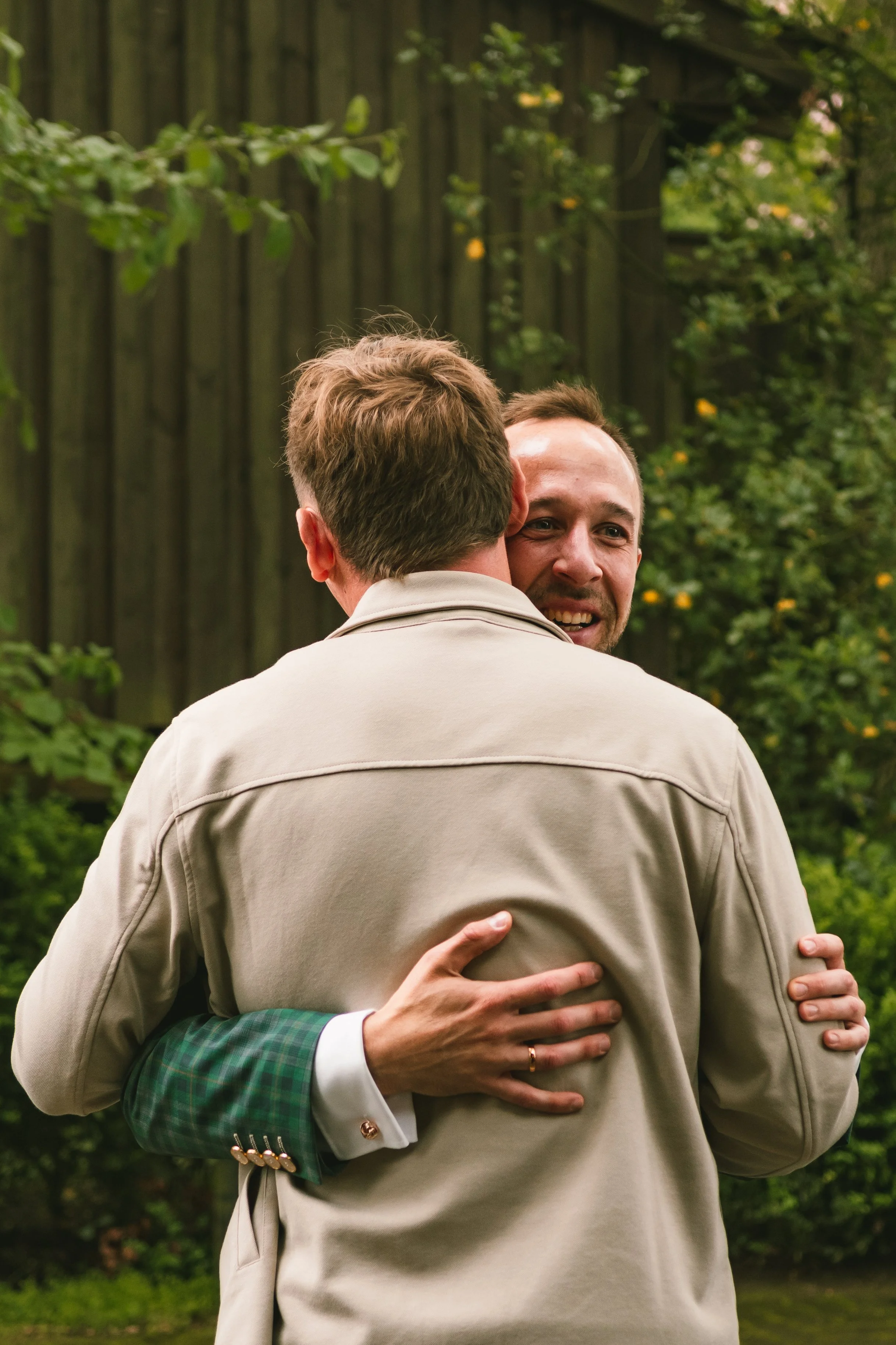 Two men hugging outdoors, seen from the back and side, with green trees and a wooden fence in the background.