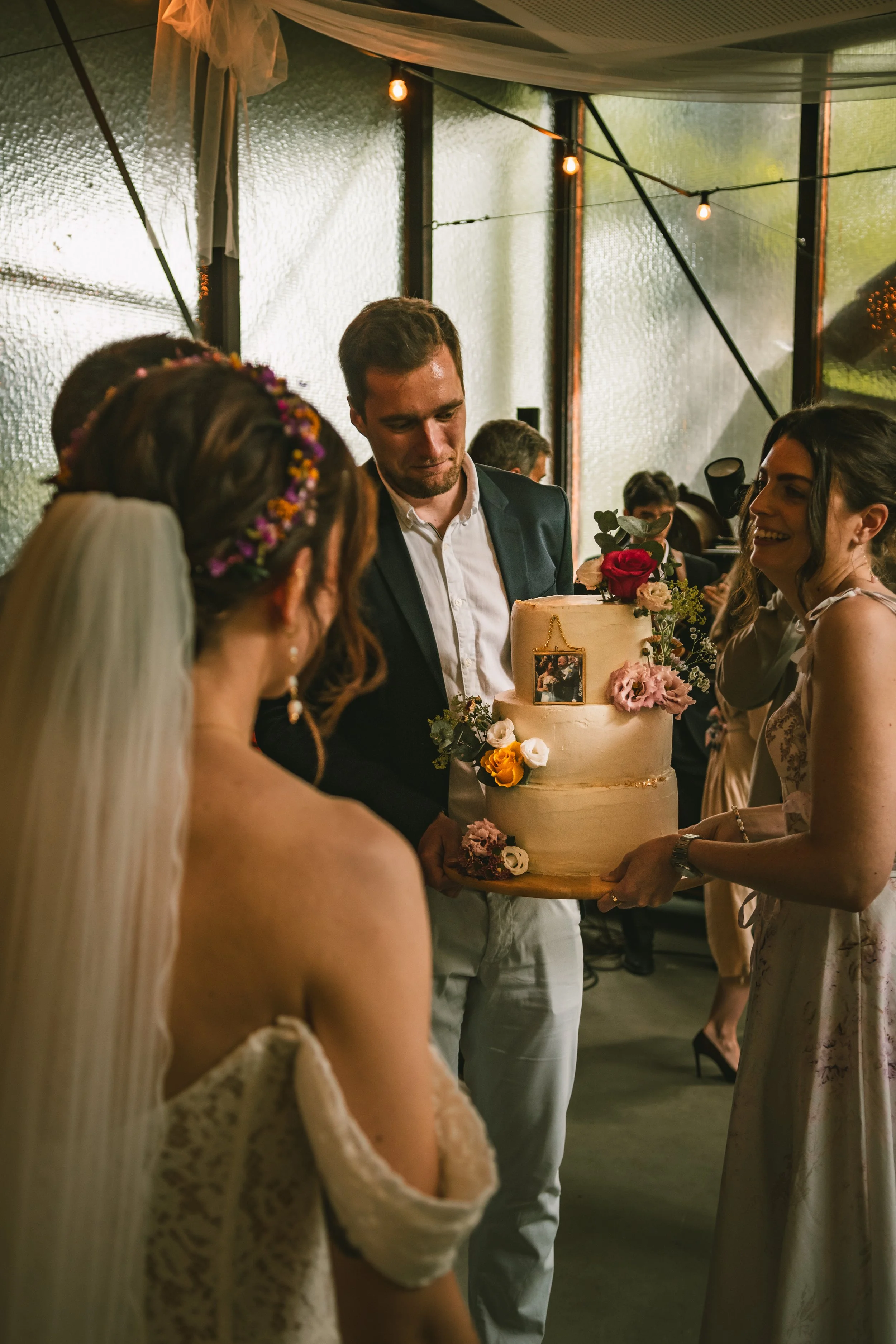 People gathered around a wedding cake at a celebration, likely a wedding reception, with a woman in a wedding dress and a man in a suit.
