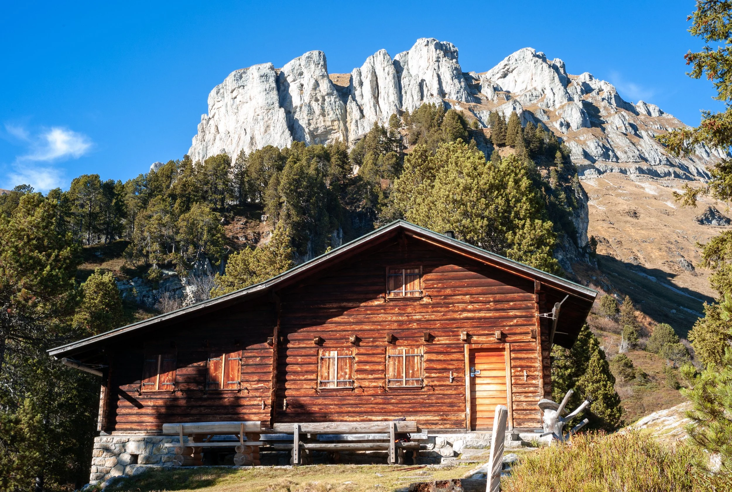 A scenic mountain landscape with a wooden cabin in the foreground, surrounded by trees and rocky peaks under a clear blue sky.