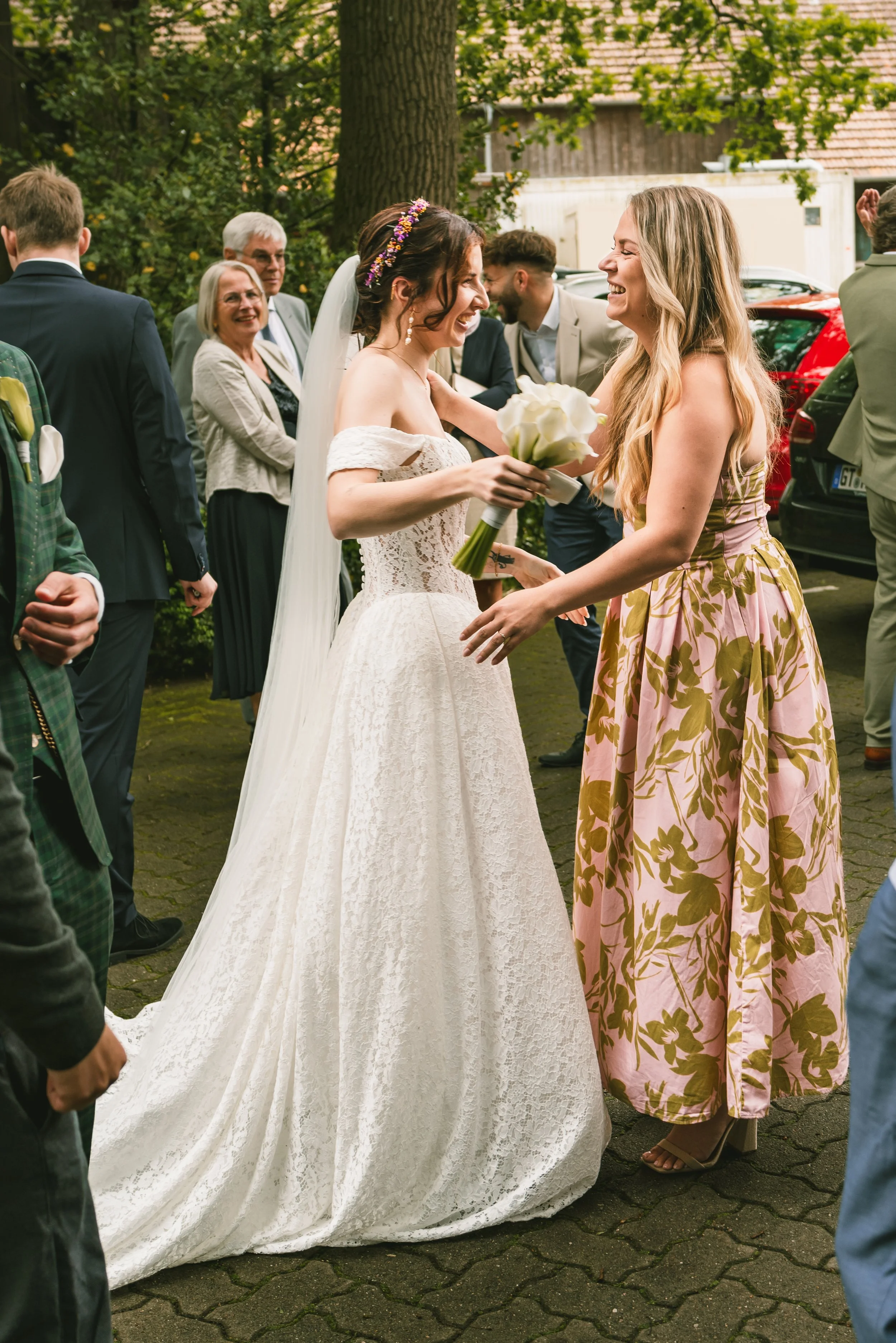 Two women, one in a wedding dress and the other in a floral dress, smiling and holding hands at a wedding celebration outdoors, surrounded by people in formal attire.