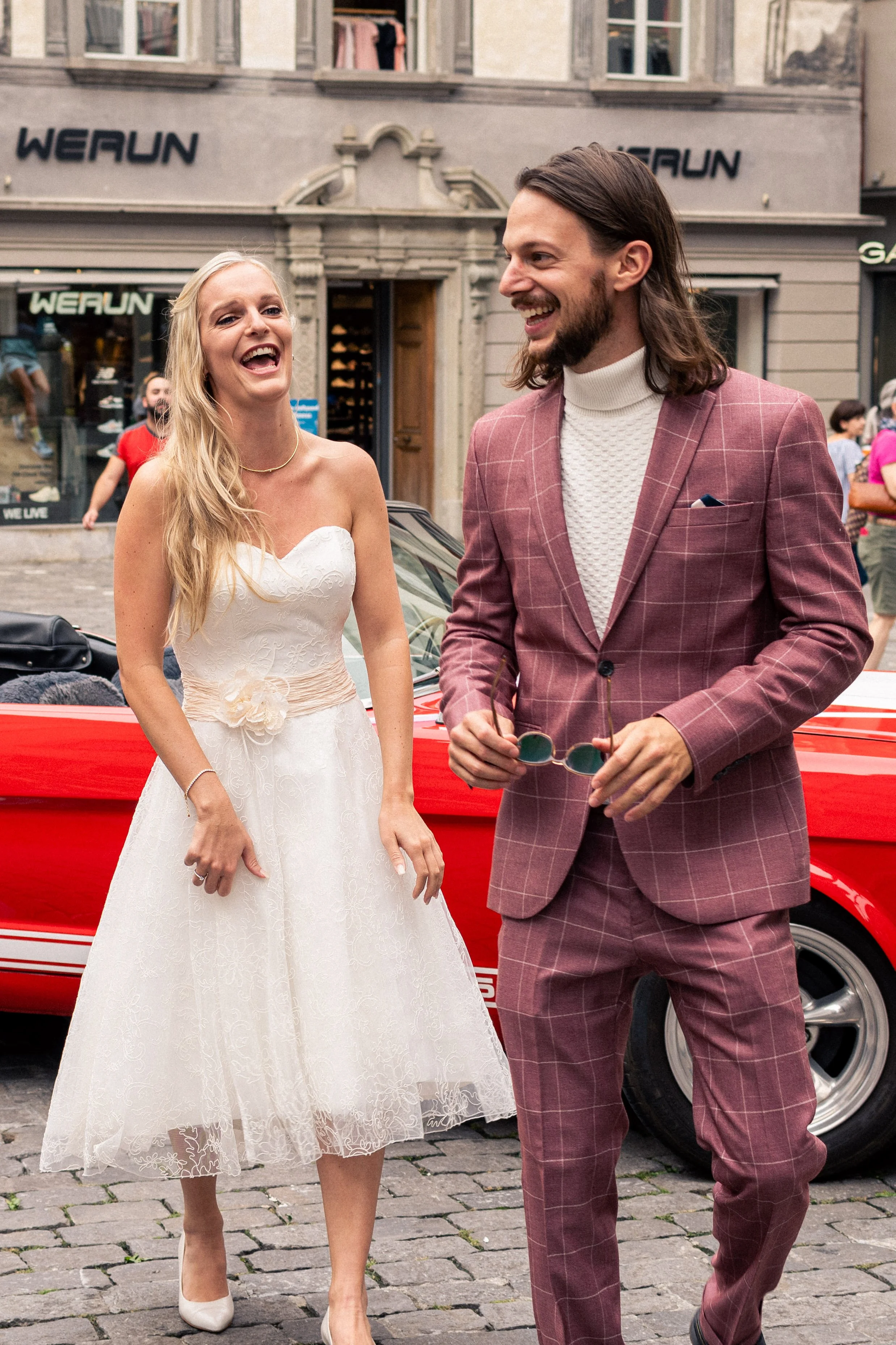A smiling woman in a white wedding dress and a smiling man in a checked suit walk together on a cobblestone street in an urban setting, with buildings and people in the background.