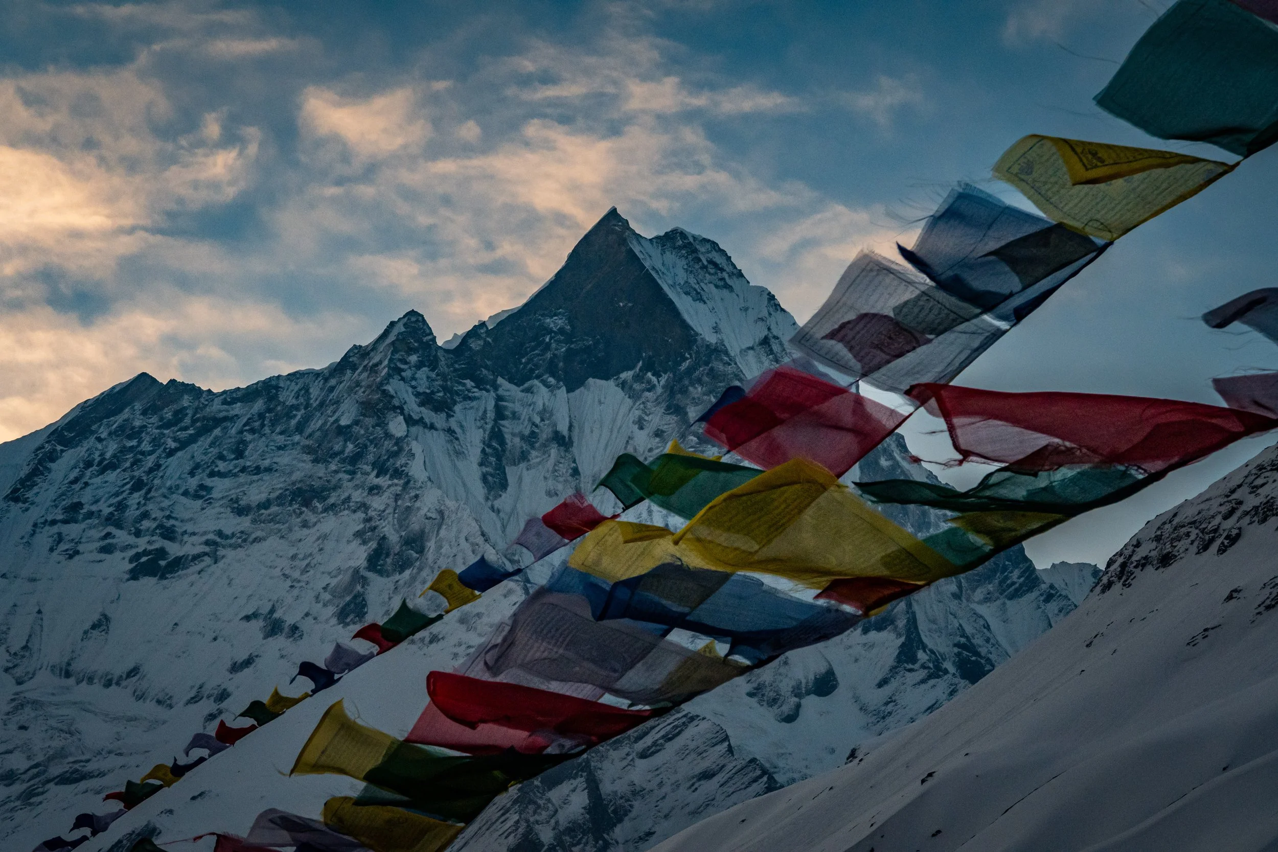 Colorful prayer flags fluttering on a snowy mountain landscape with a peak in the background and a partly cloudy sky.