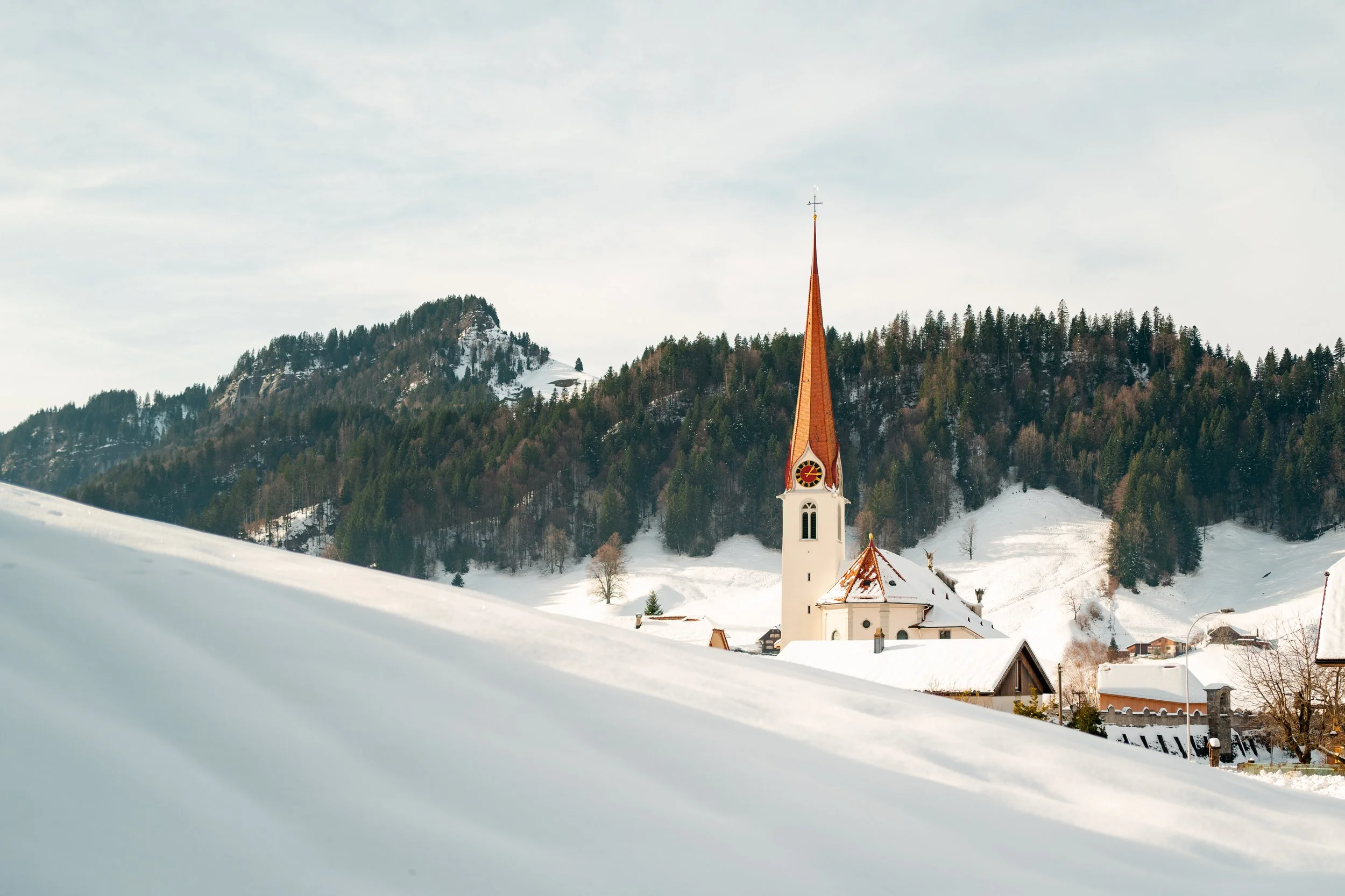 A snow-covered landscape with a church featuring a tall, pointed steeple with a clock and cross on top. There are mountains and trees in the background under a cloudy sky.