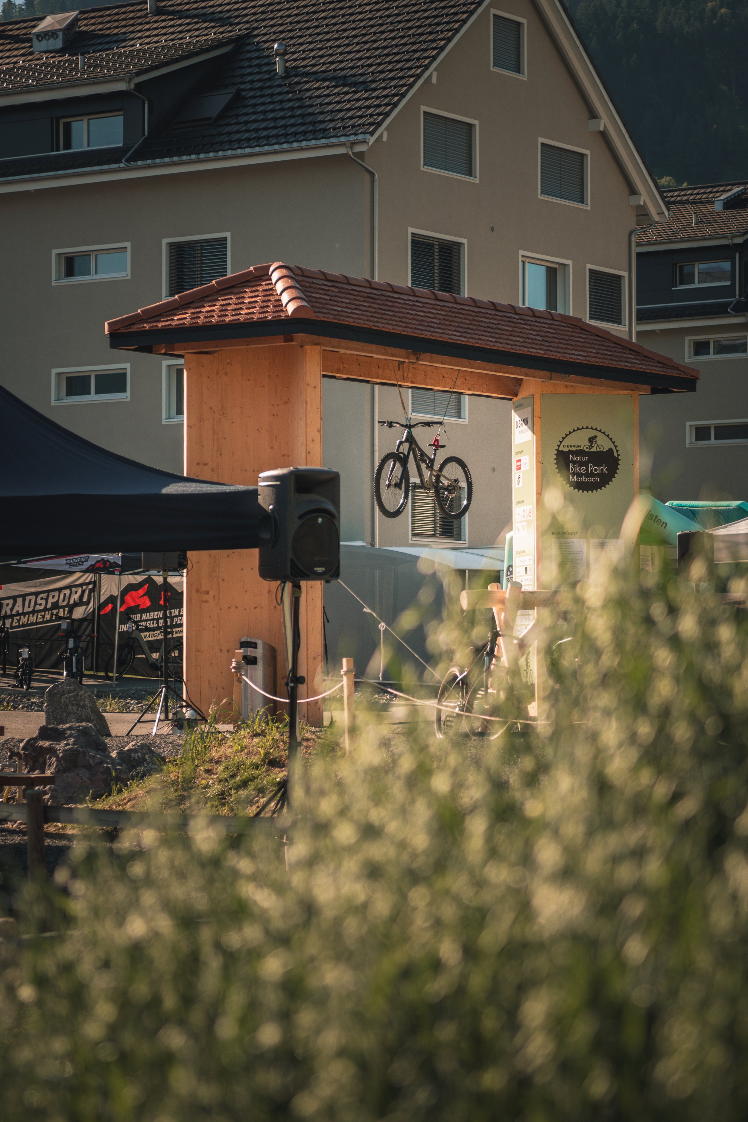 A wooden shelter with a red tiled roof, a bicycle hanging from the ceiling, and a sign that reads 'Natur Bike Park Marbach' in front of a beige residential building with multiple windows.