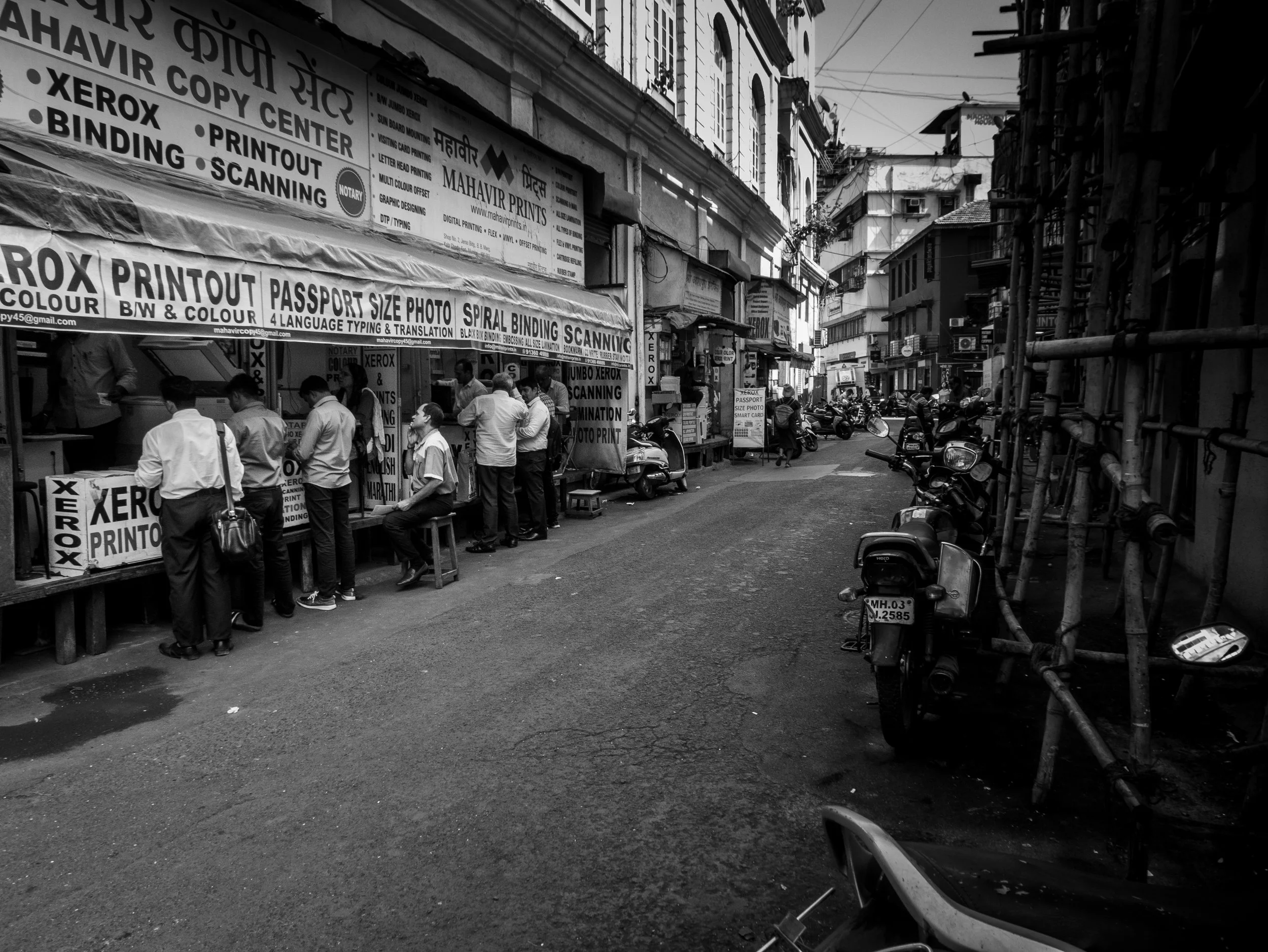 Street scene in an urban area with a print shop storefront, motorcycles parked along the street, and people standing outside the shop. The scene is in black and white.