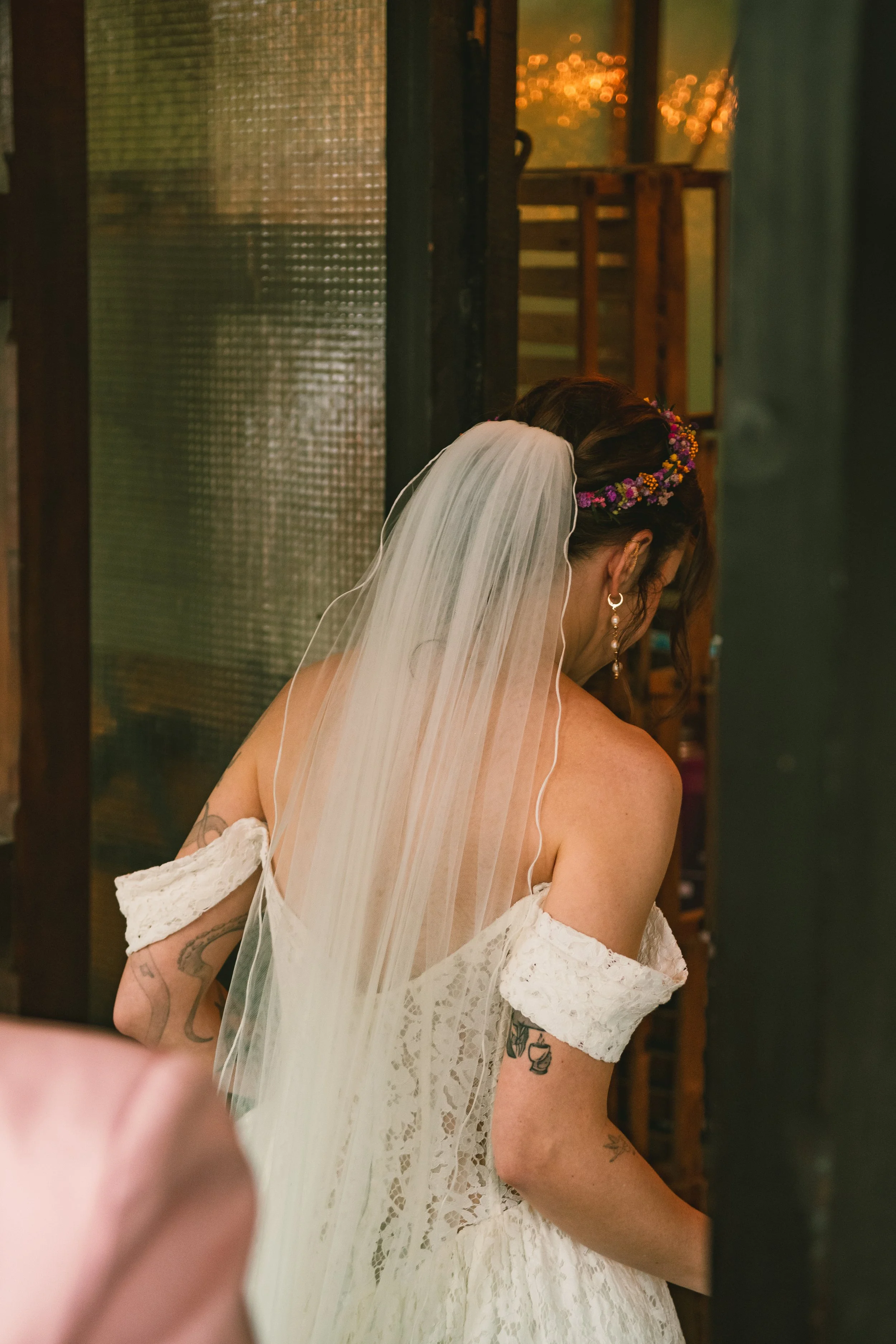 A bride on her wedding day wearing a white lace off-shoulder wedding dress, a veil, and a floral headpiece, with tattoos visible on her arm, standing indoors with a blurred background.