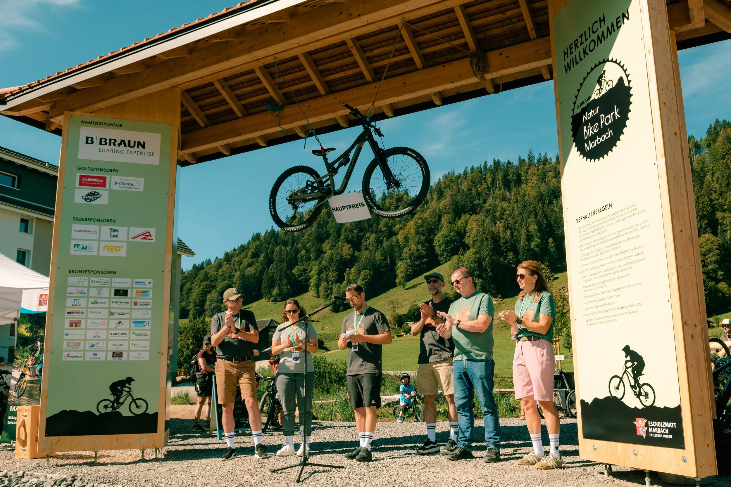 Group of people gathered at a bike park entrance with large signs displaying sponsors, a bicycle hanging from the wooden structure, and a scenic mountain background on a sunny day.