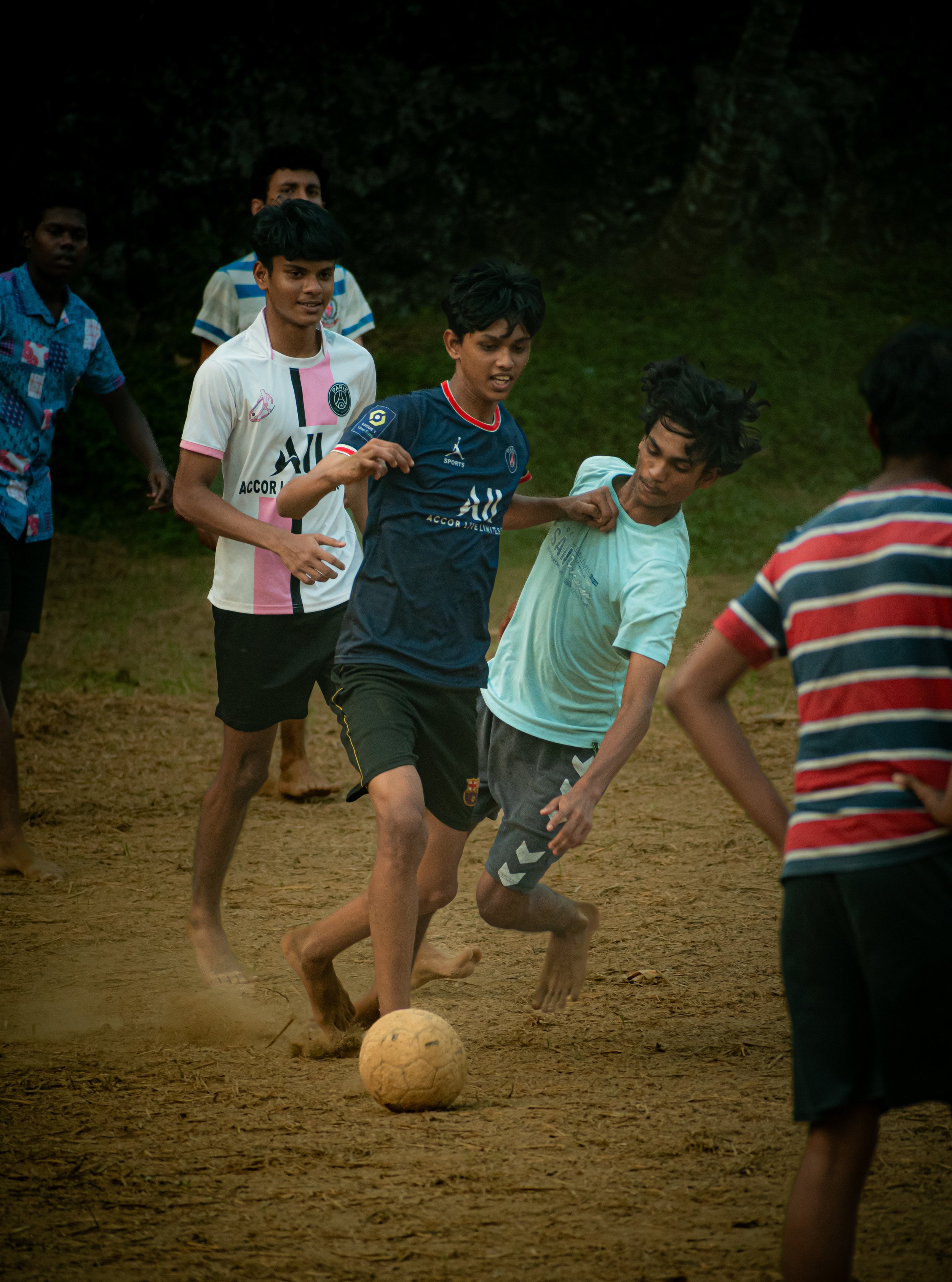 Young boys playing soccer barefoot on a dirt field, with some other children watching.