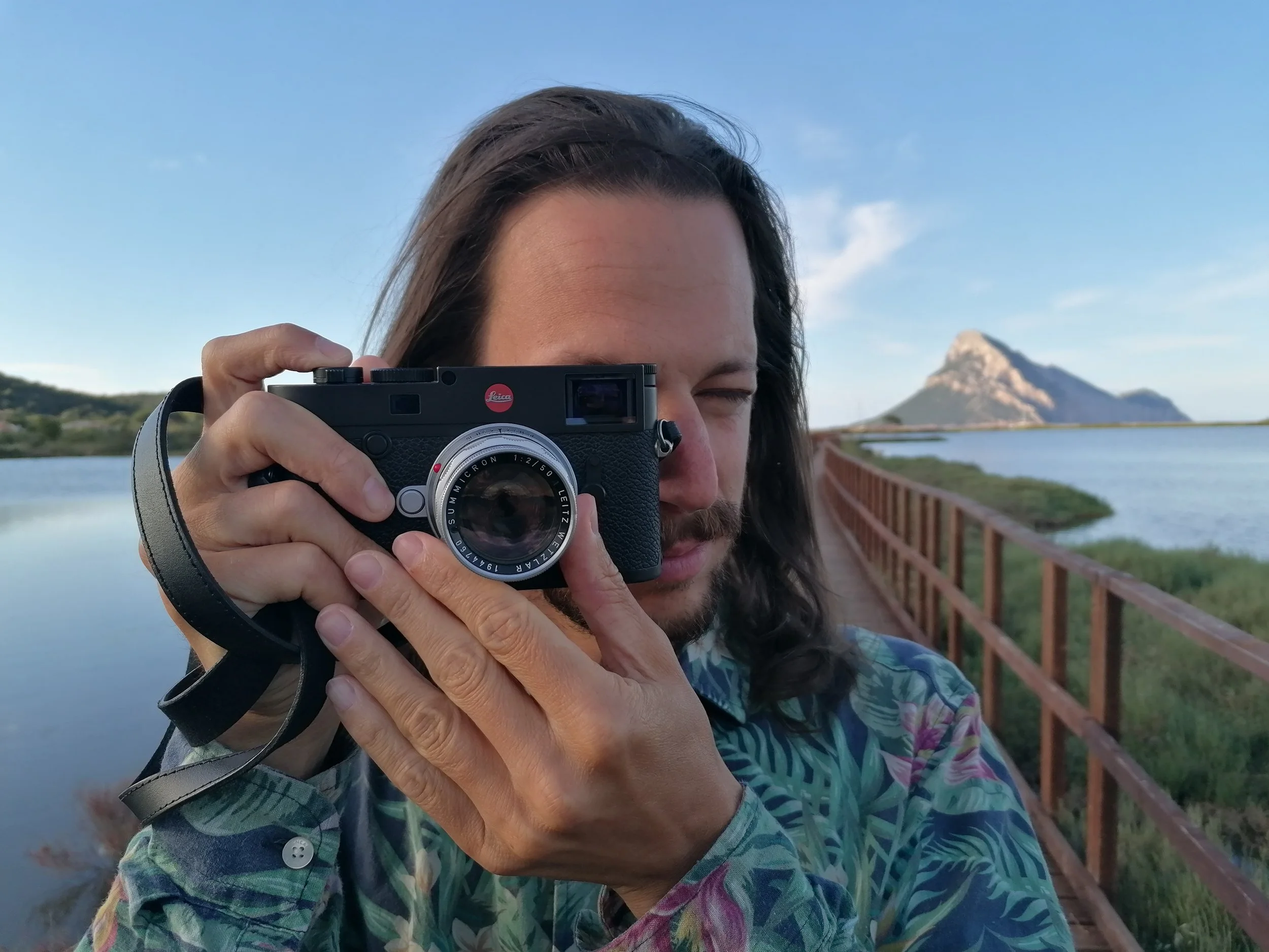 A person with long hair and a beard is taking a photo with a black camera that has a red logo, standing on a wooden bridge over water with mountains and a blue sky in the background.