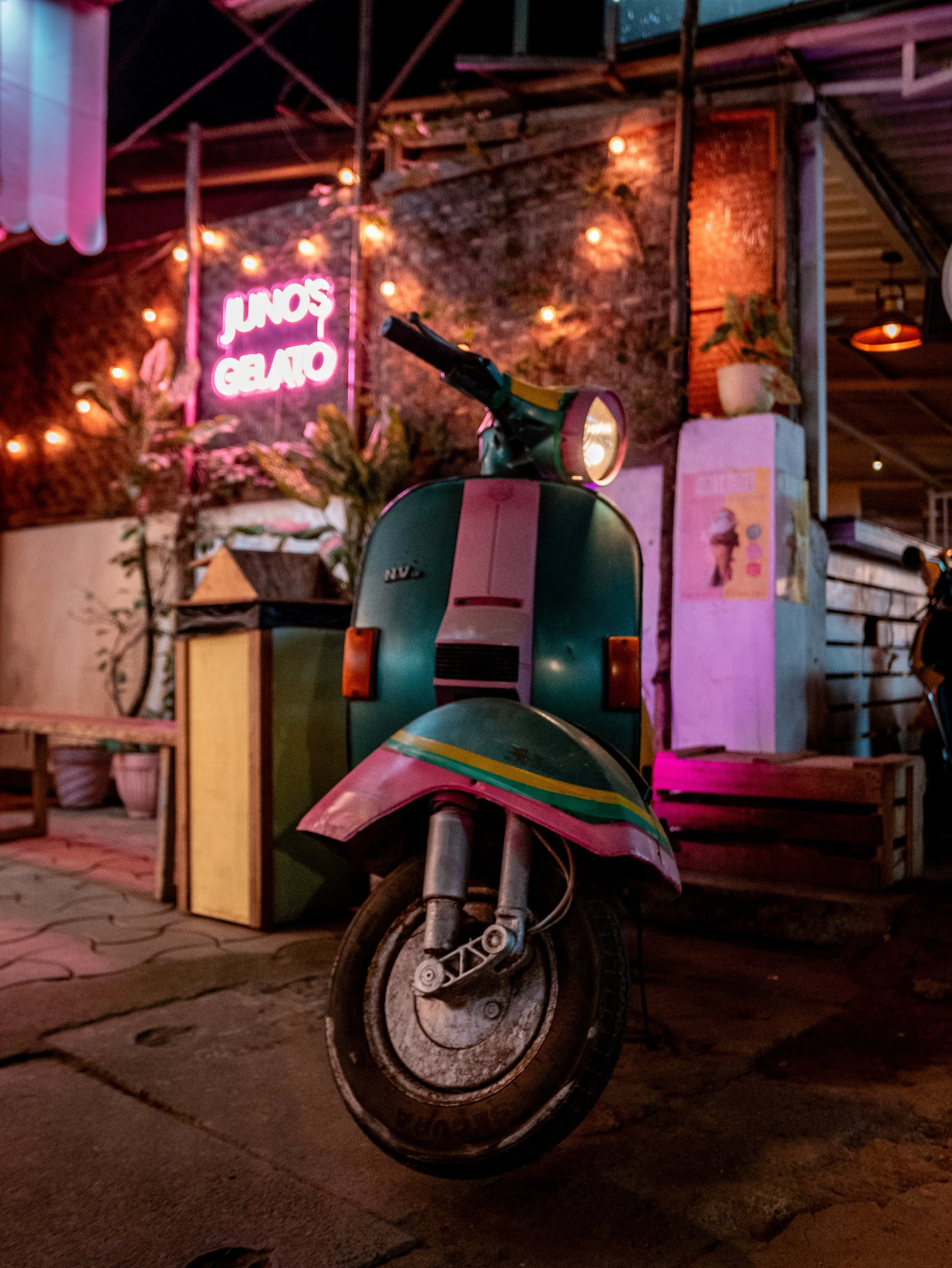 A colorful scooter parked on a brick floor under warm indoor lighting. In the background, there's a brick wall with guitar string lights hanging, a neon sign in pink that reads 'JUNOS GELATO,' and various plants and furniture.