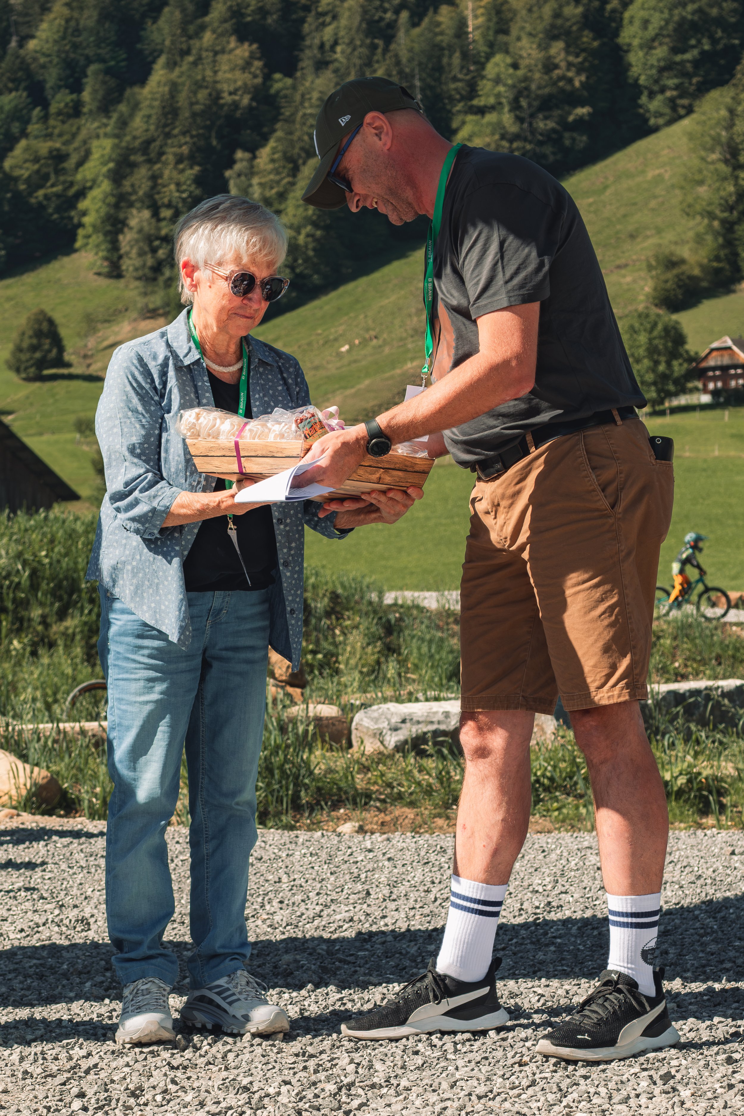 An older woman and a young man are outdoors on a gravel path with green hills and trees in the background. The woman is holding a gift basket, and the man is standing with his head bent, wearing athletic clothes and socks, and taking the gift basket.