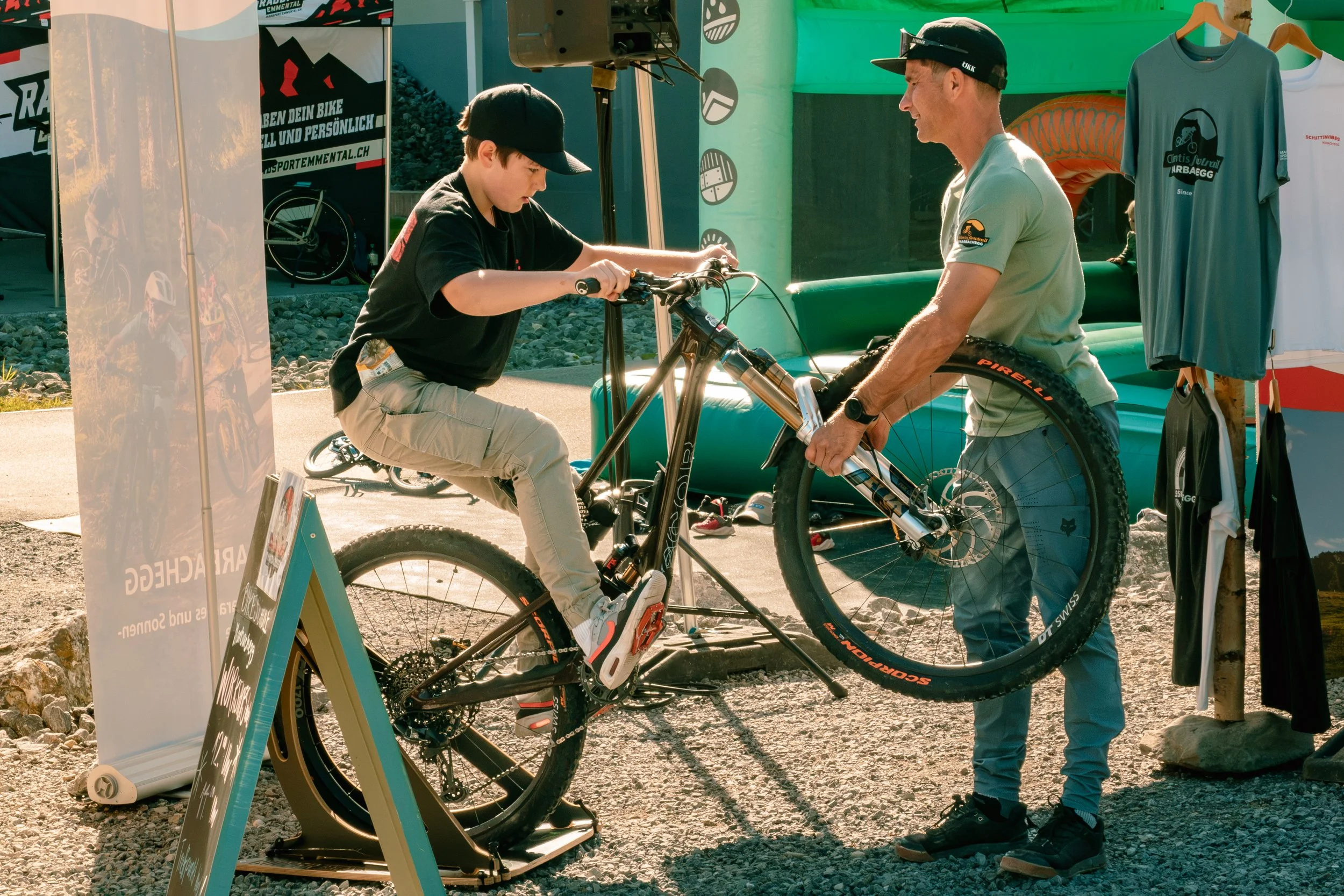 A boy practicing mountain biking on a stationary trainer at an outdoor event, with an adult man assisting him. There are mountain biking shirts hanging for display nearby, and promotional banners in the background.