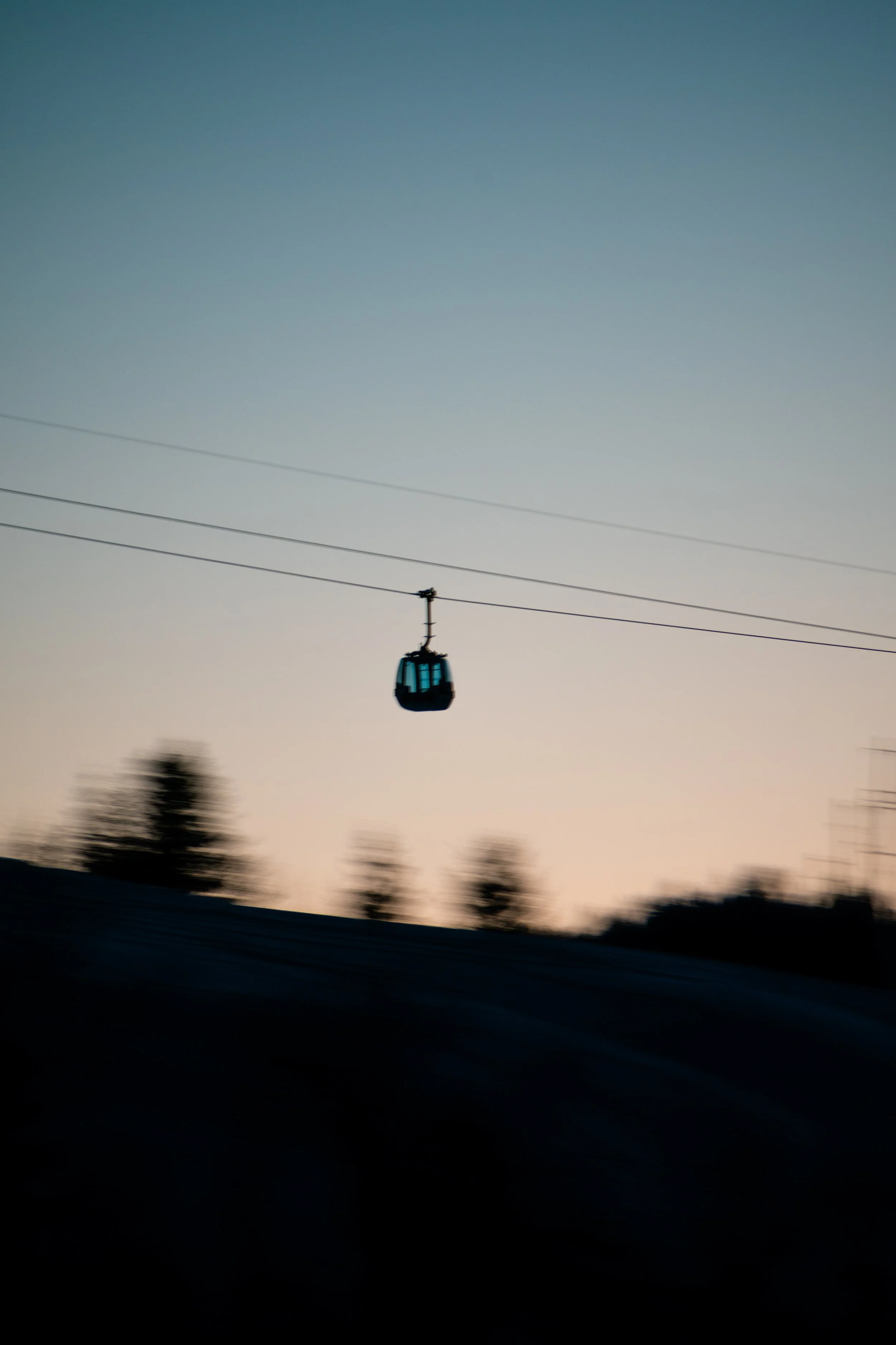 A cable car suspended in the air against a blurry sunset sky with trees at the bottom.