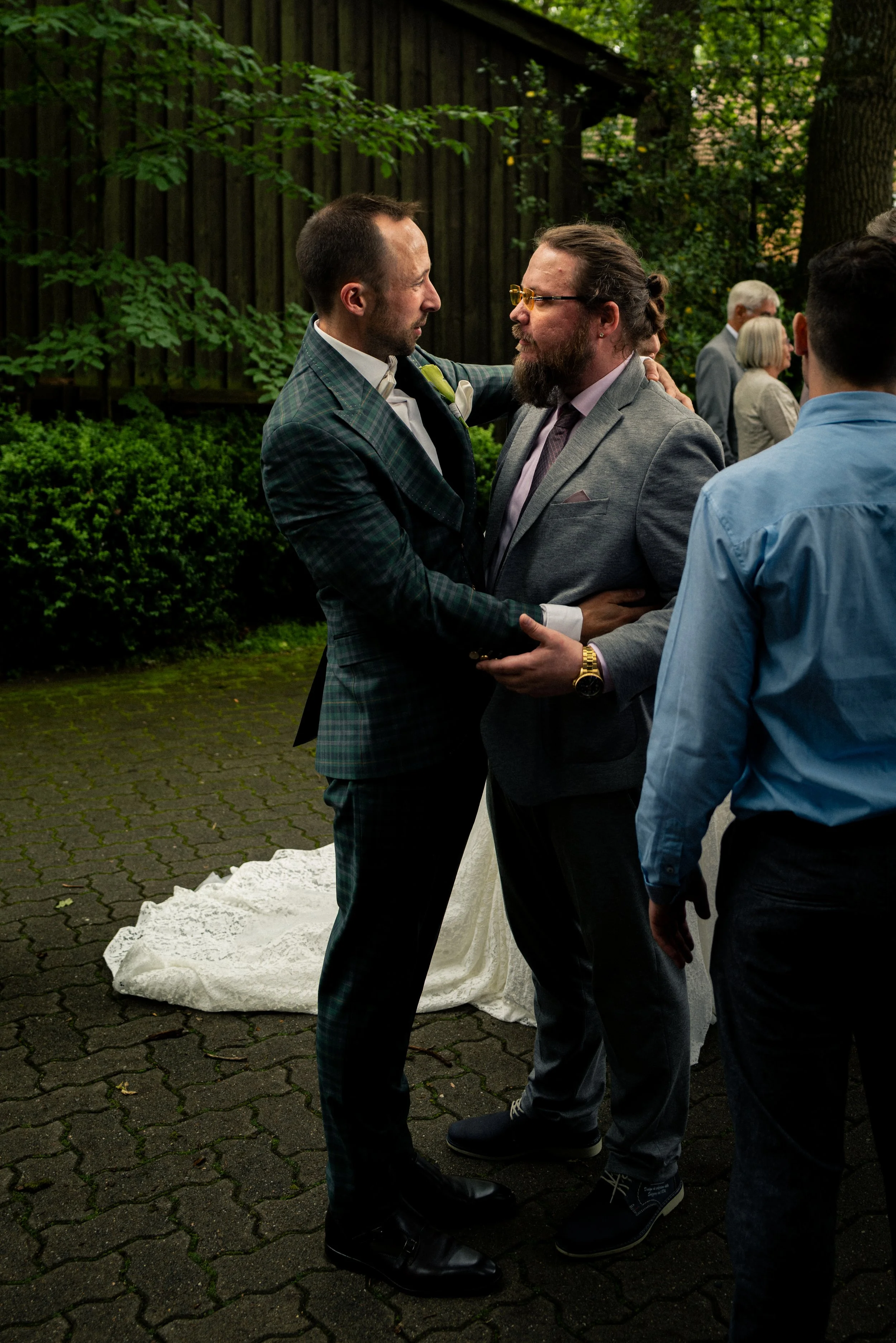 Two men dressed in suits are embracing at an outdoor event, possibly a wedding, with others in the background. They are standing on a paved area with greenery and trees behind them.