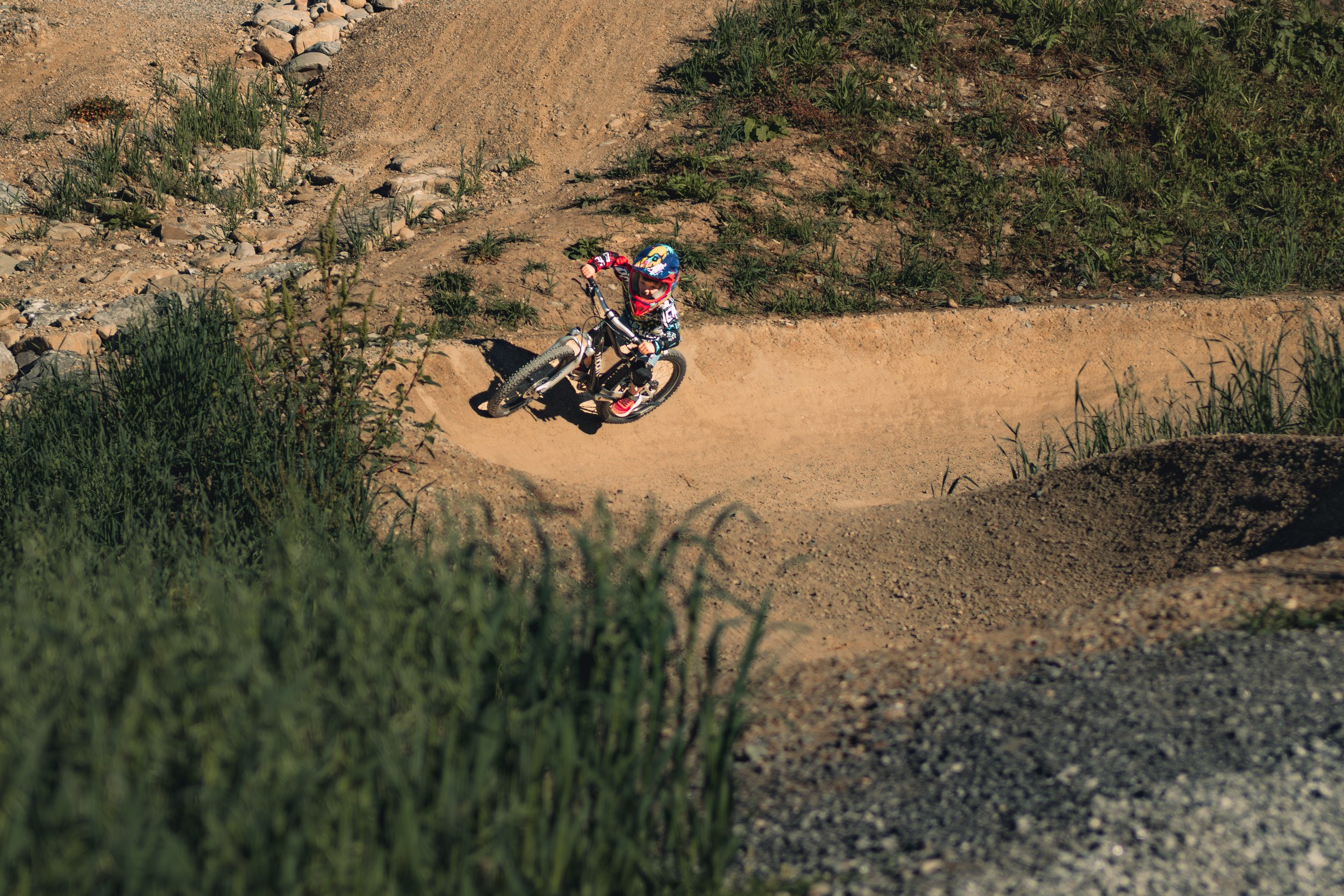A young child riding a mountain bike on a dirt trail in a natural outdoor setting with patches of grass and rocks.