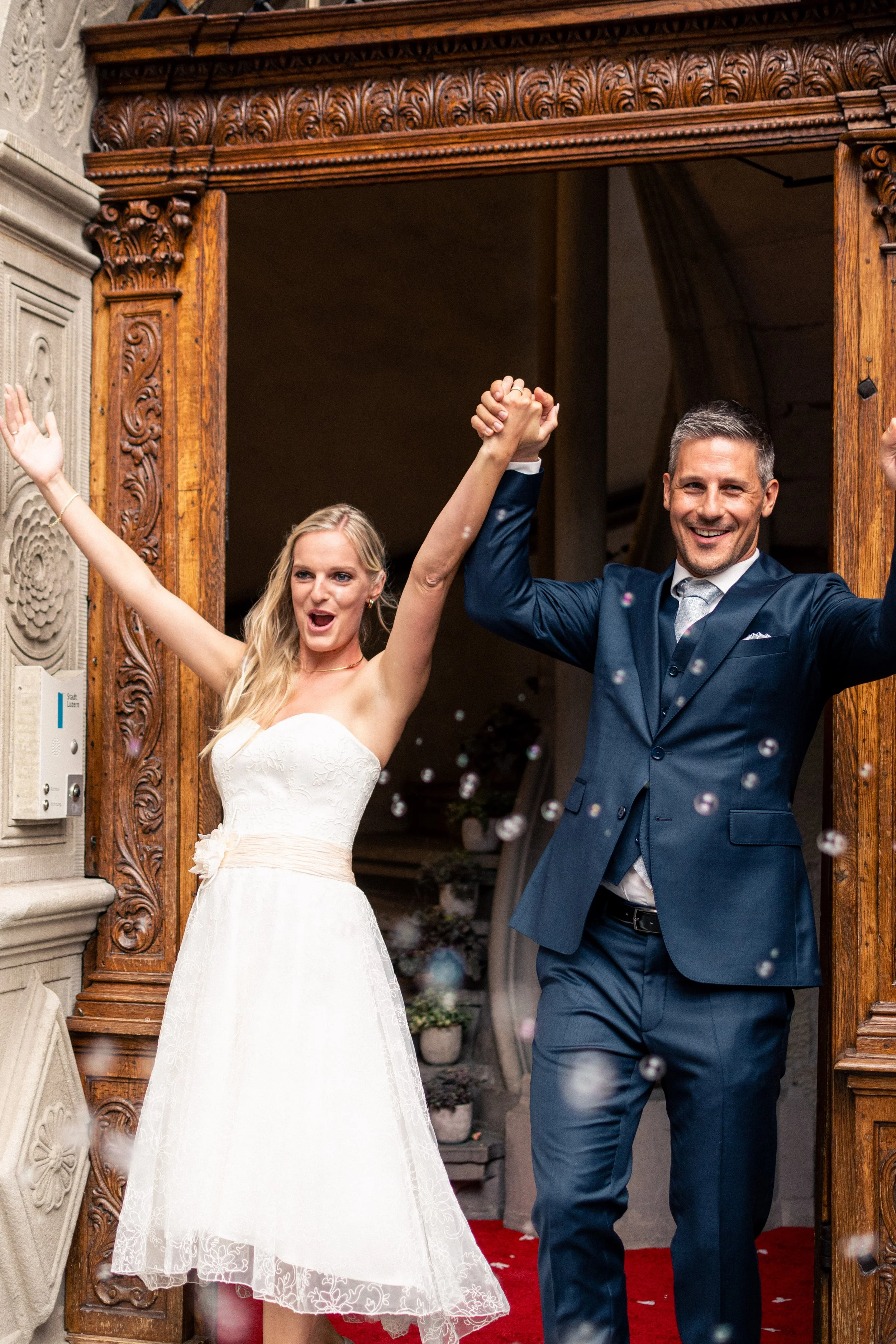 A bride and groom celebrating their wedding, holding hands, smiling, and raising their arms in joy as they exit a building with a fireplace and intricate wooden carvings, surrounded by falling confetti.