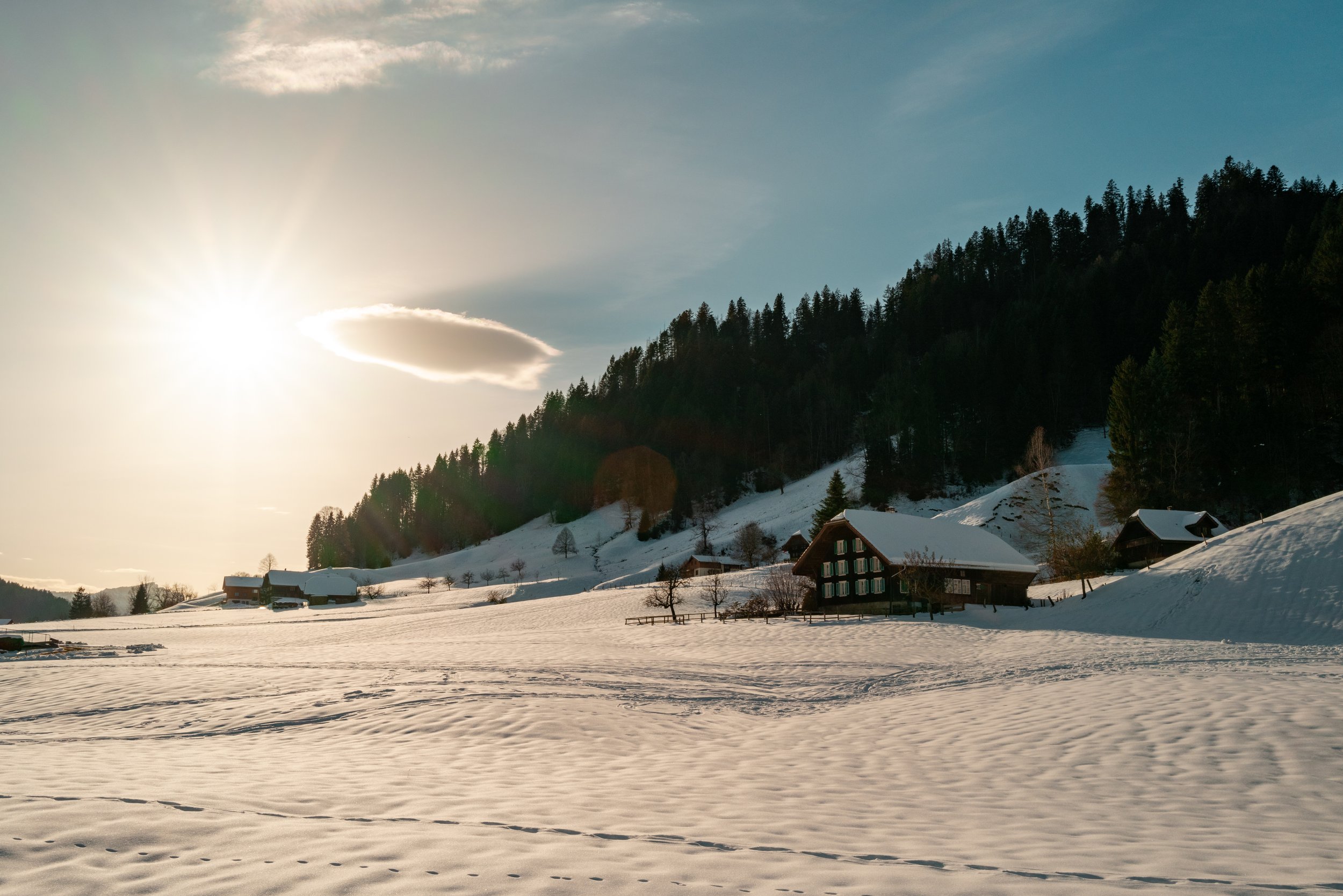 Snow-covered landscape with a few houses, pine trees on a hillside, and the sun partially obscured by clouds, casting a glow over the scene.