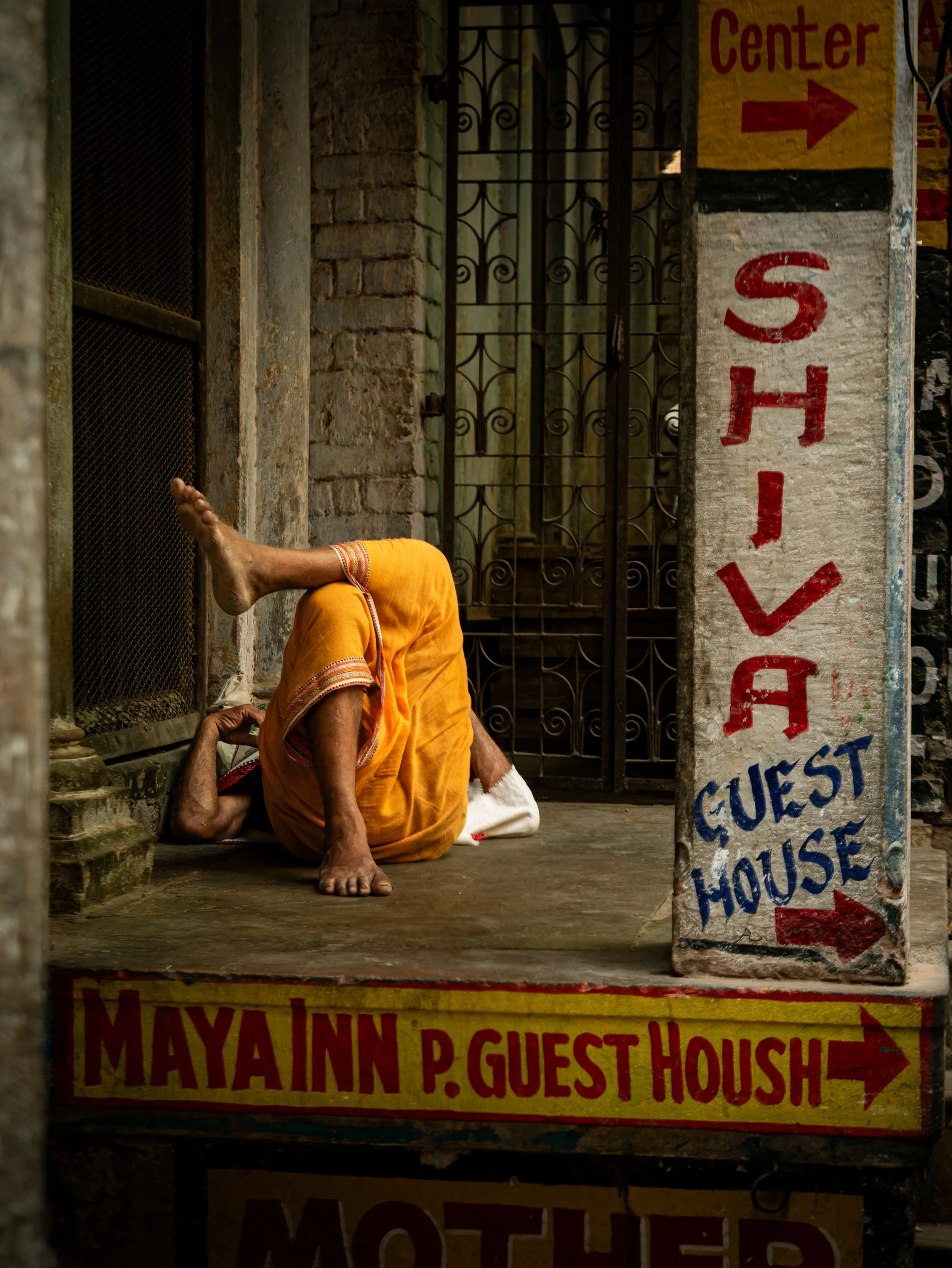 A person sitting on the ground wearing a yellow outfit, with his head resting on his knees and his arm stretched out on the floor. The person appears to be in a shaded part of an alley with brick walls and painted signs, including one that reads 'MAY