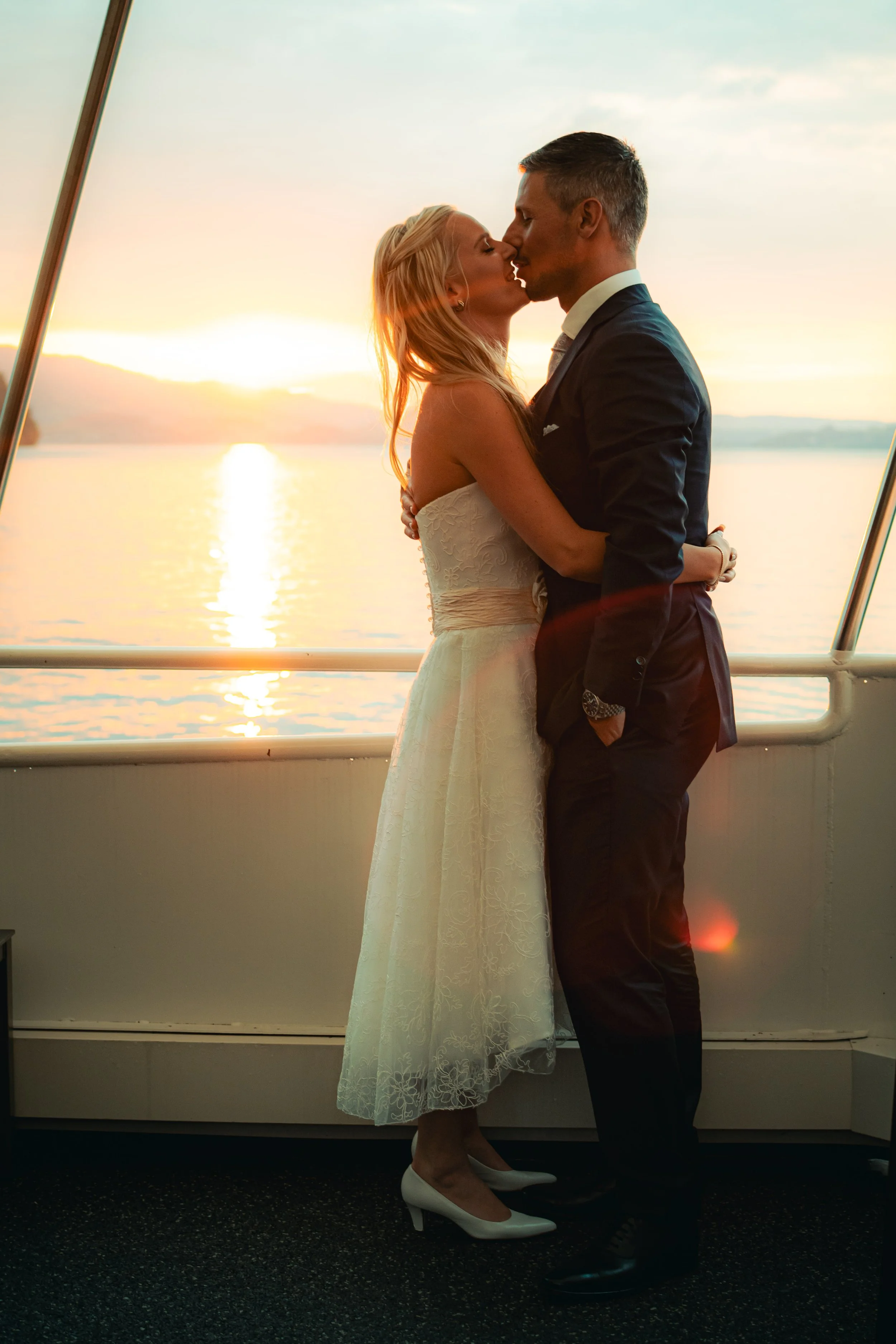 A couple kissing on a boat during sunset, with water and mountains in the background.