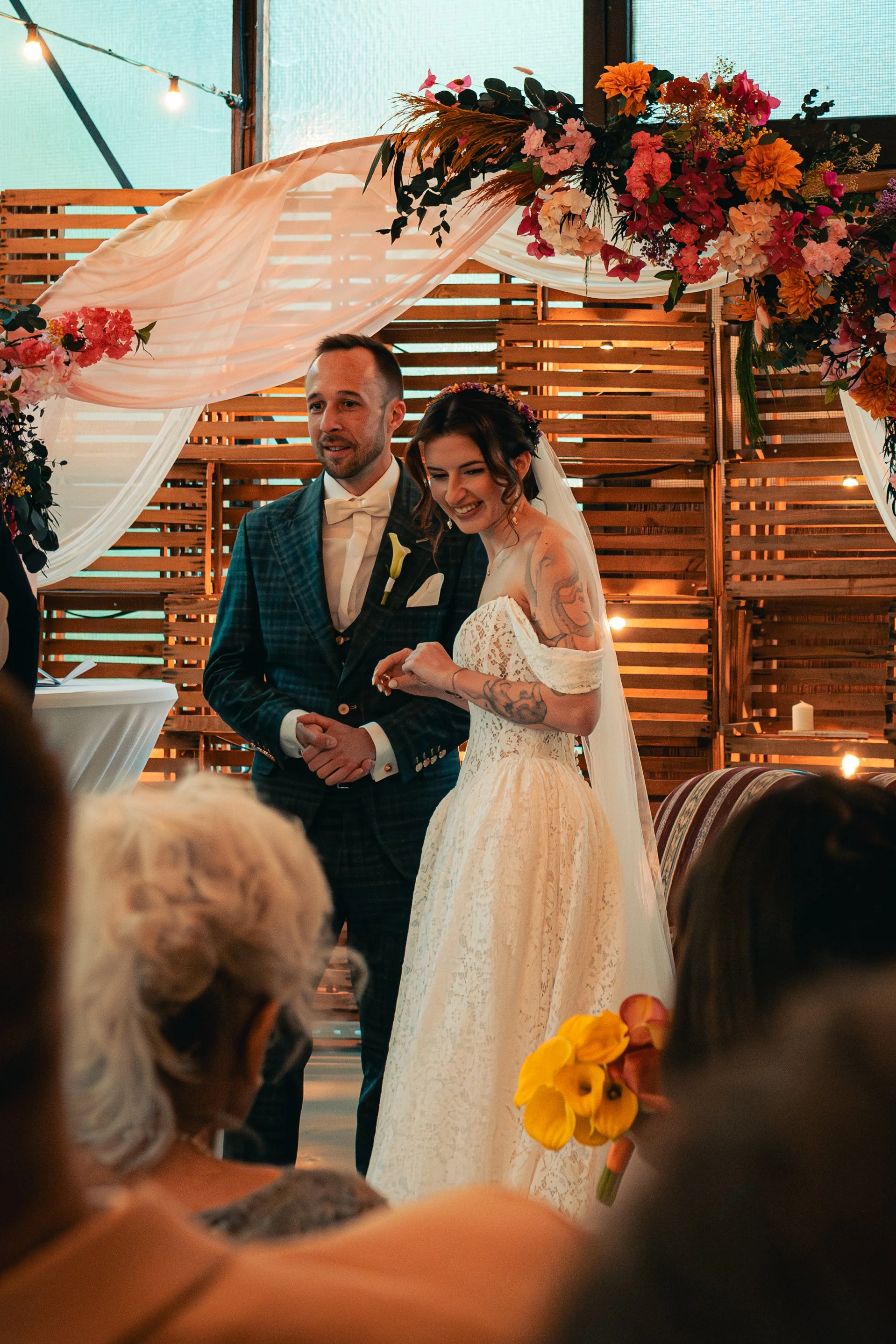A couple, possibly bride and groom, standing under a floral arch at a wedding ceremony. The bride is smiling and holding a bouquet of yellow flowers, while the groom is dressed in a dark plaid suit with a bow tie. Guests are visible in the foreground, and the setting is decorated with string lights and wooden panels.