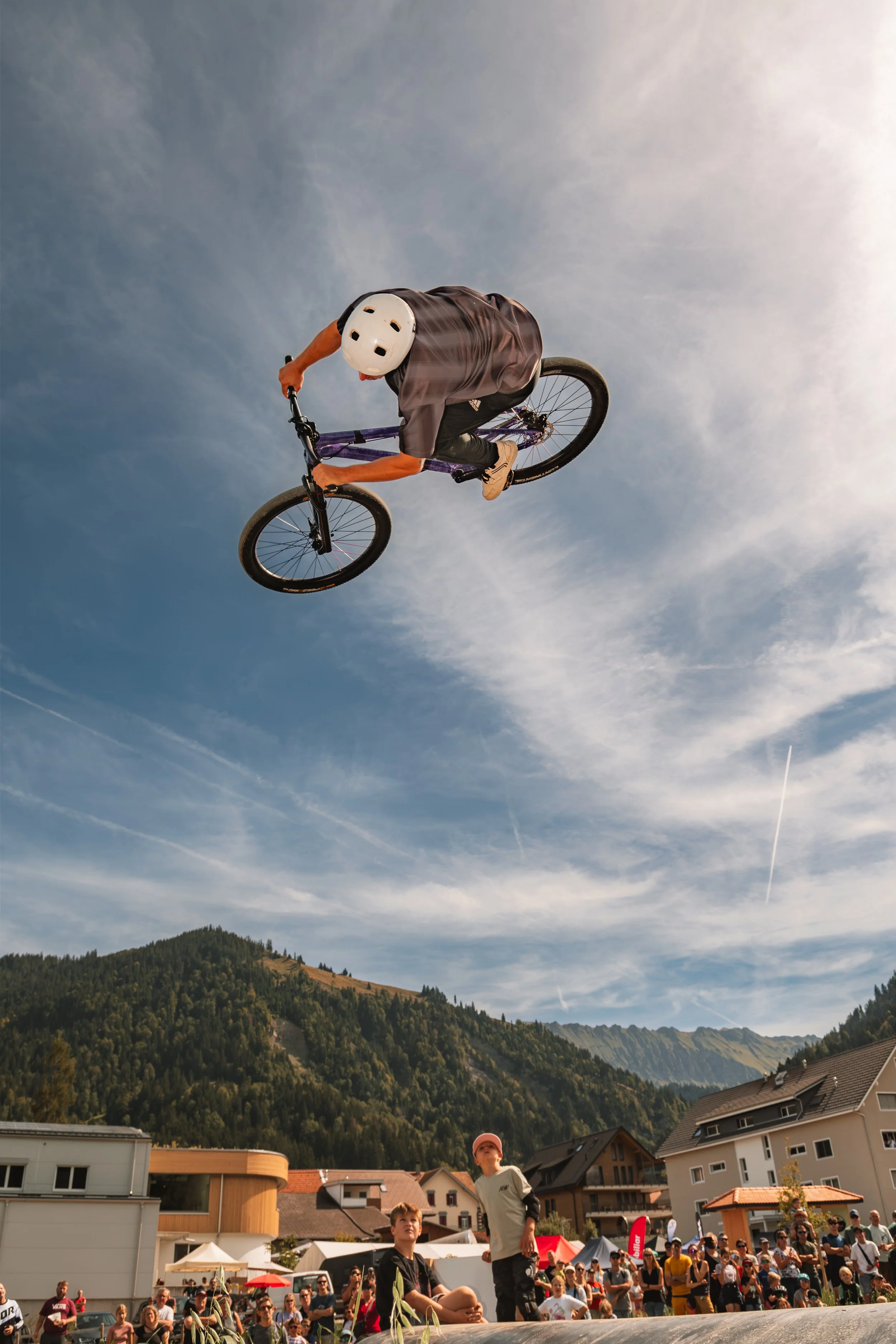 A BMX rider performing a trick in mid-air during a public event, with spectators watching in a small town with mountains in the background on a clear day.