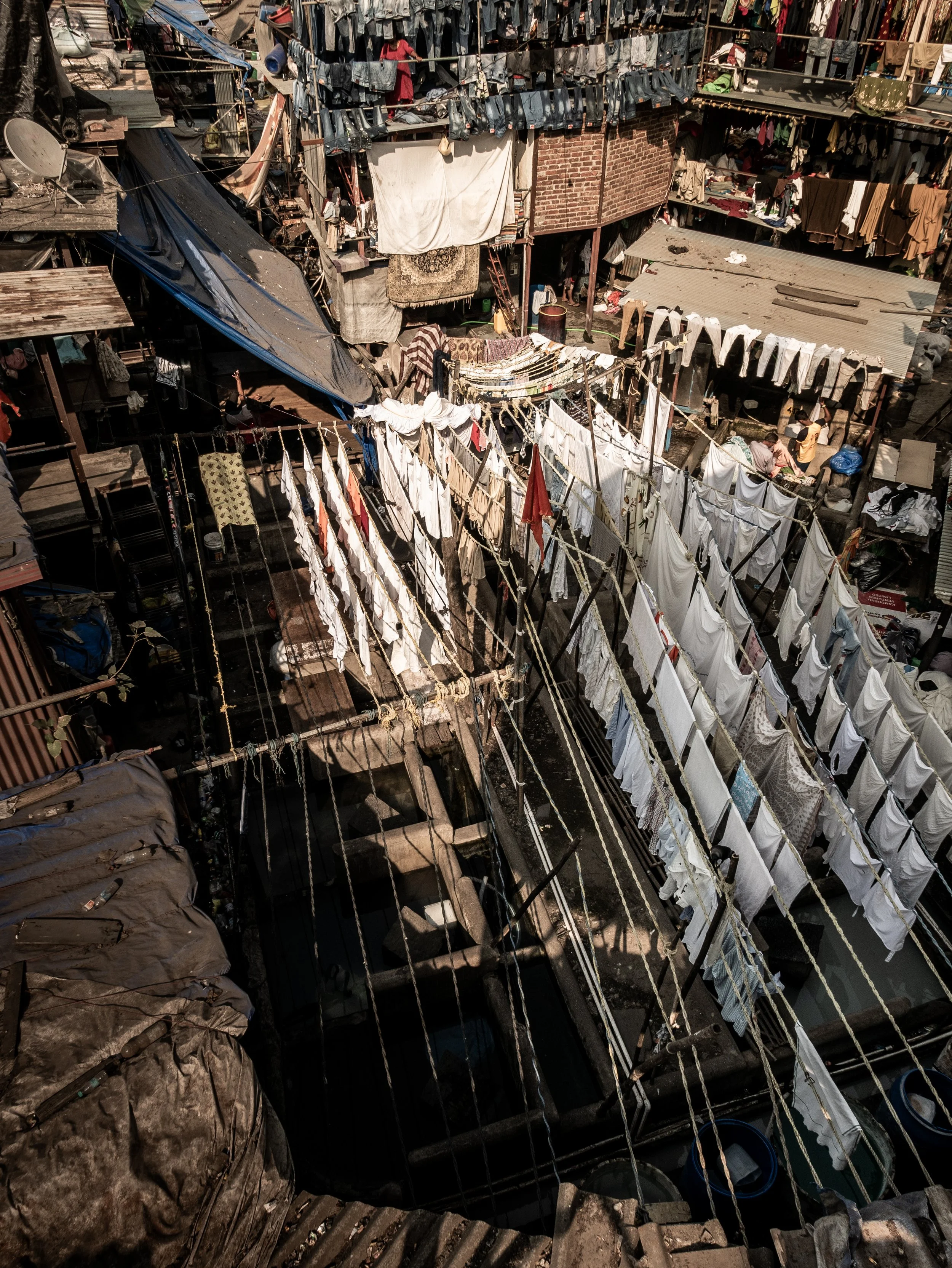 An outdoor laundry area with clothes hanging on lines to dry, multiple levels of laundry drying racks, and clothing items scattered around. The setting appears to be in a densely populated residential area with makeshift structures and brick walls.