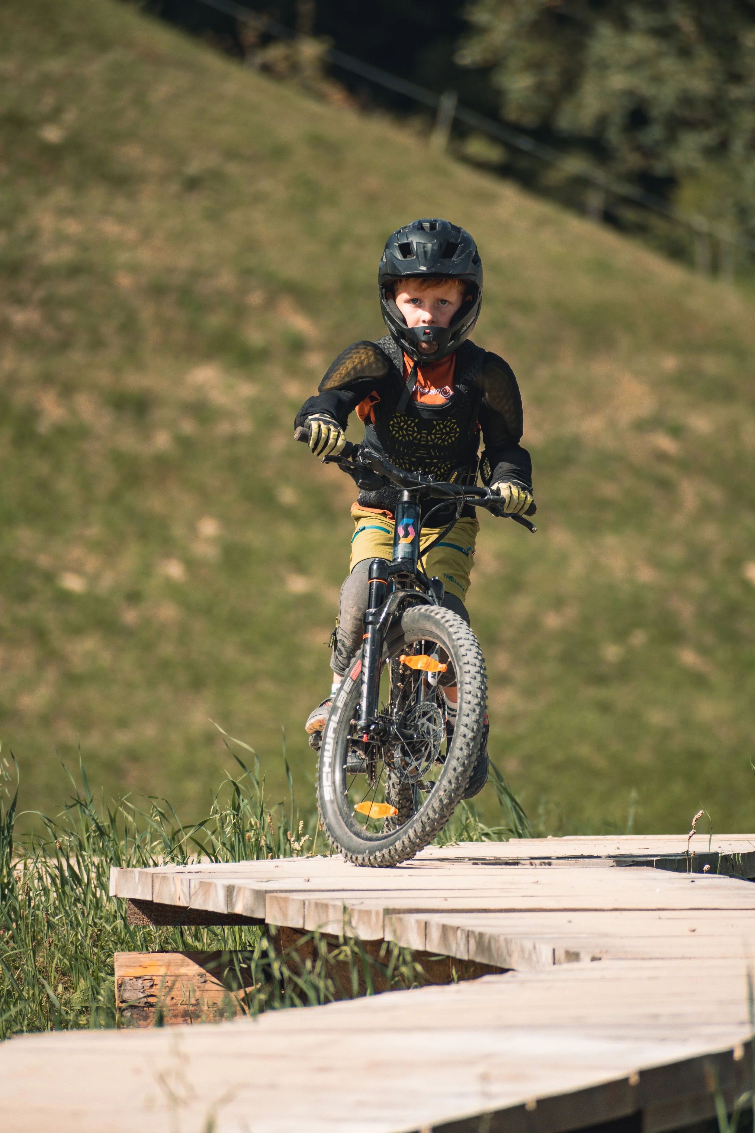 A young boy wearing a black helmet and protective gear riding a mountain bike on a wooden platform in a grassy outdoor area with trees in the background.