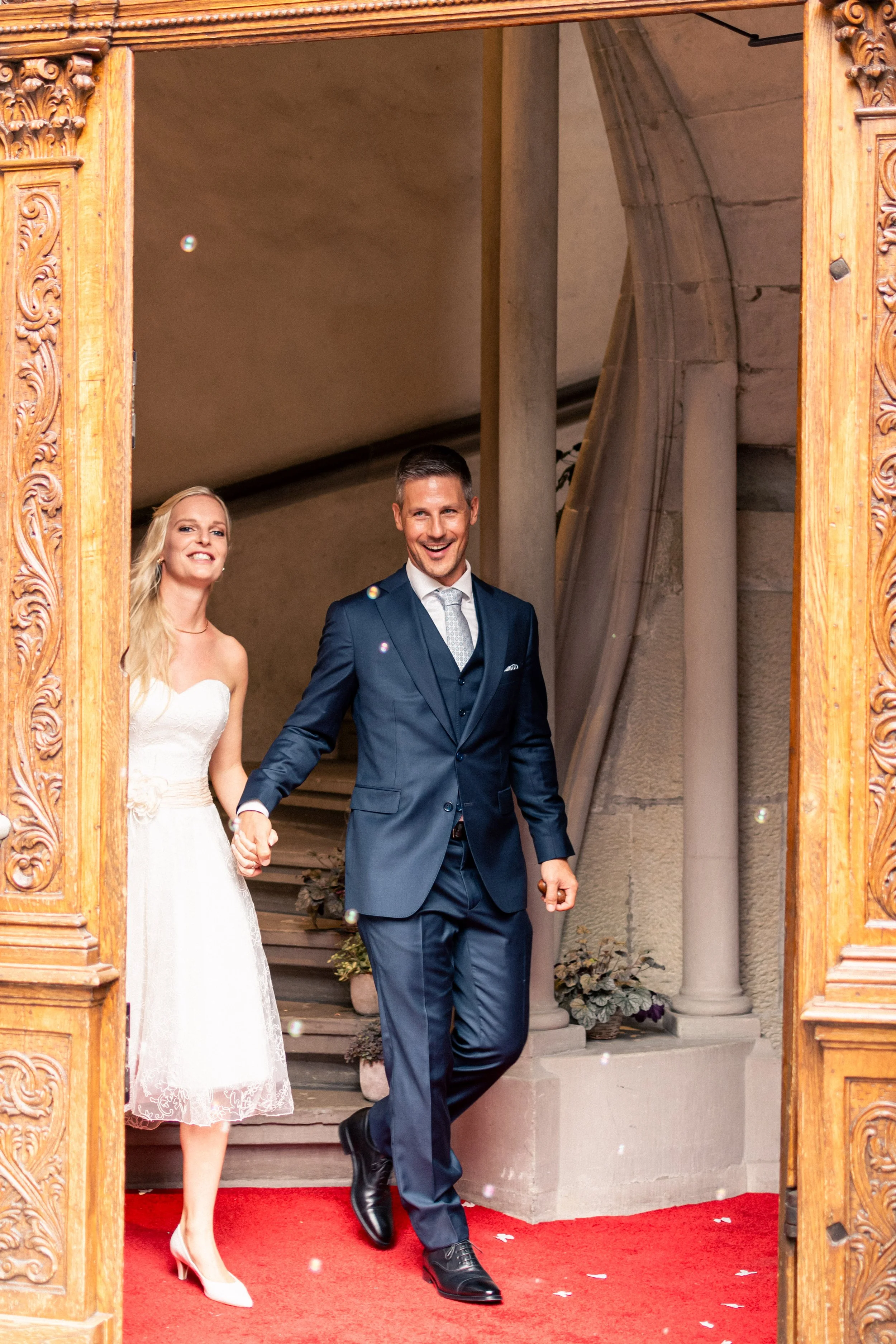 A bride and groom holding hands and smiling as they leave a building, standing under a decorative wooden archway. The bride wears a white strapless wedding dress and high heels, while the groom wears a dark blue suit with a light tie.