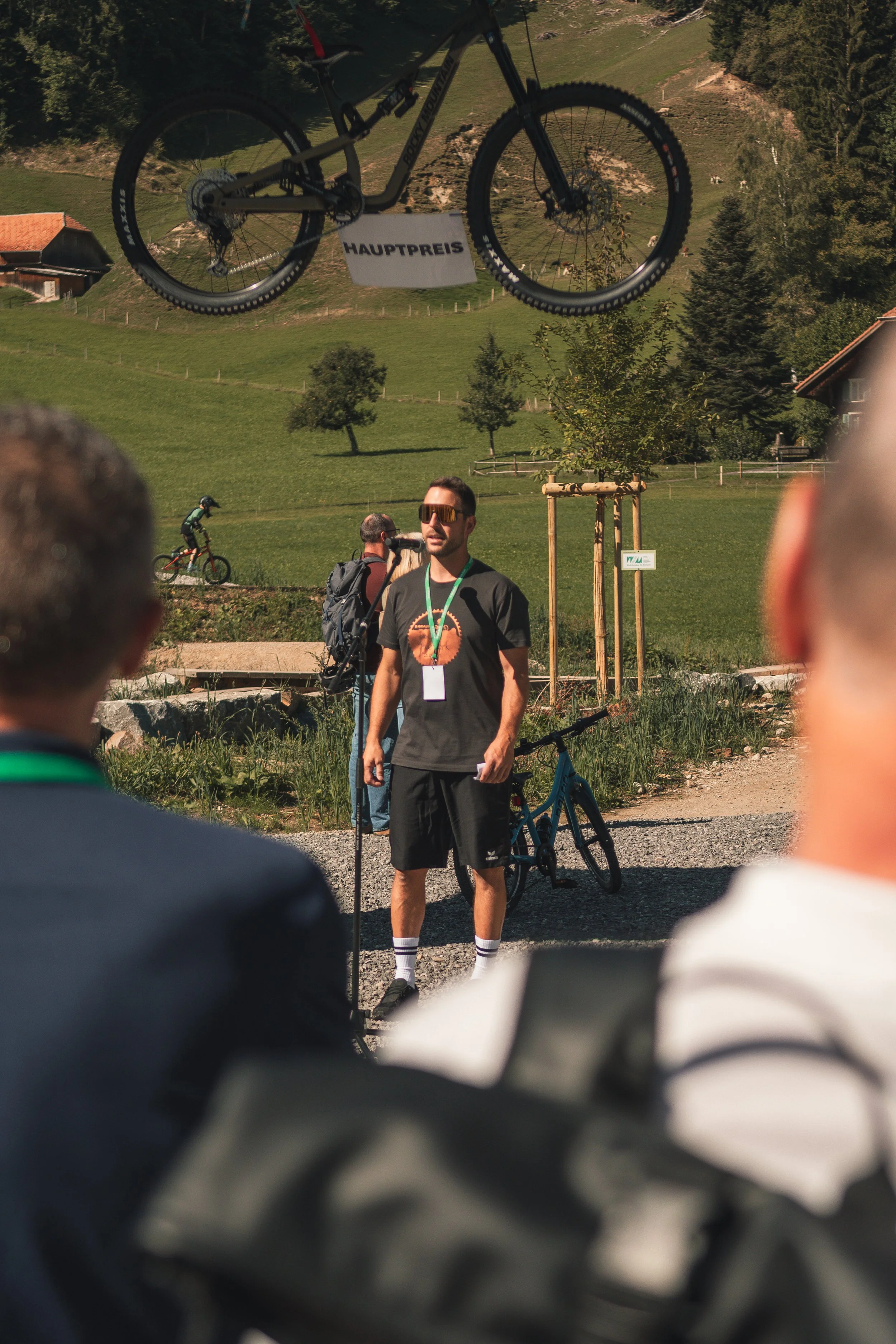 A man wearing sunglasses, a dark t-shirt, and shorts is standing outdoors in front of a group, with a bicycle beside him. Behind him, a small crowd watches, and there are green hills, trees, and a few houses in the background. The image is taken thro