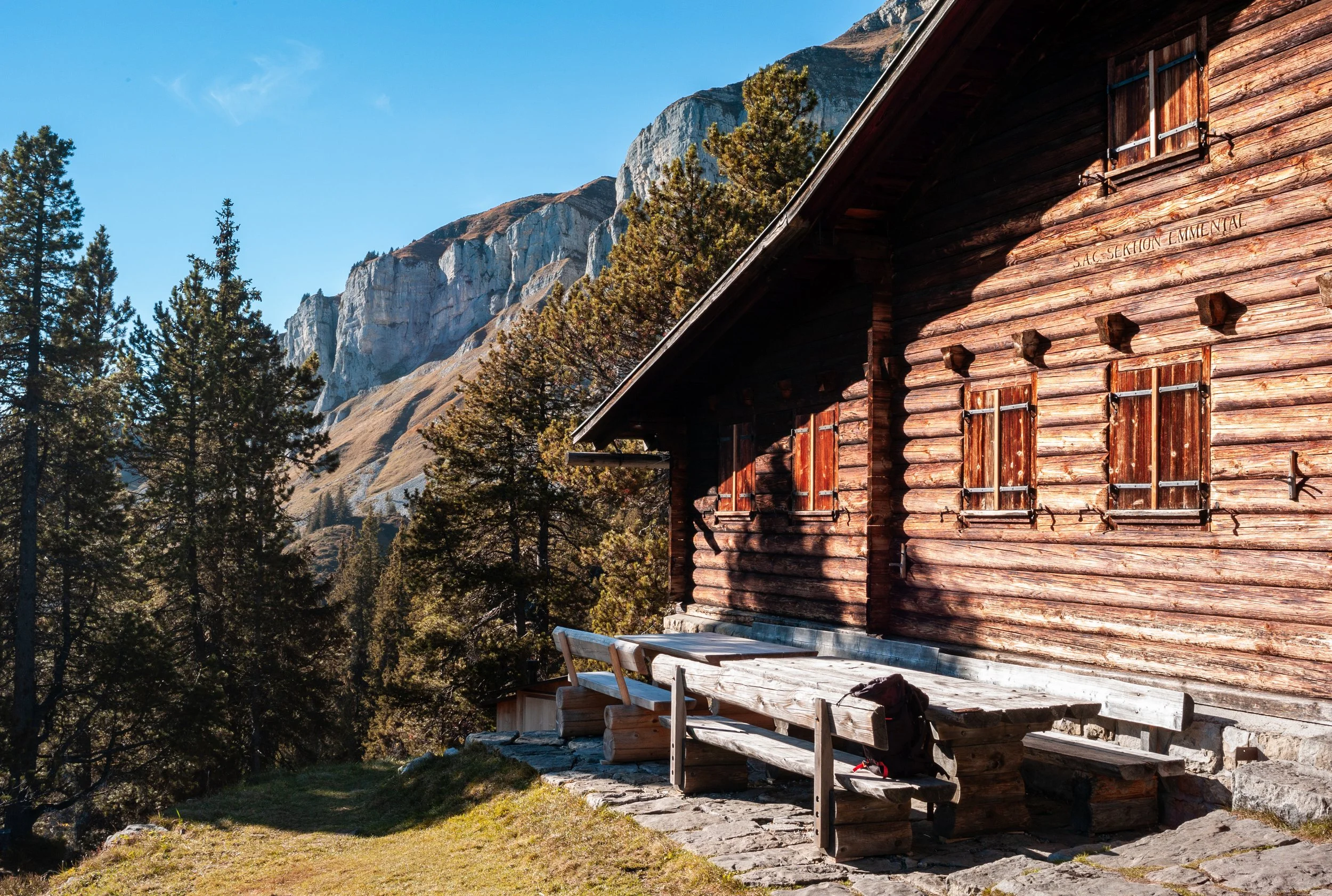 A wooden cabin in a mountain forest setting with trees and rocky mountain cliffs in the background, under a clear blue sky.