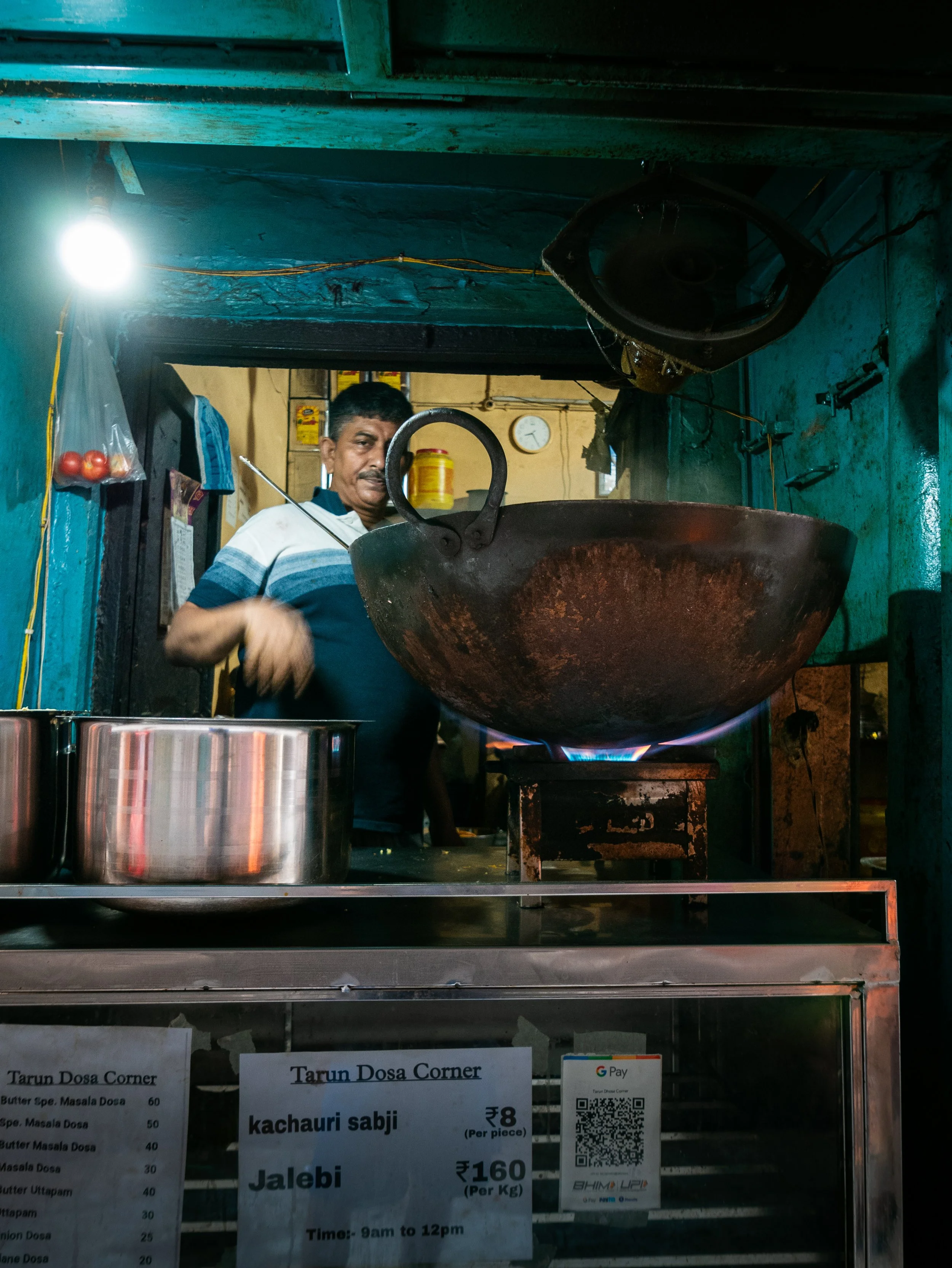 A man stands behind a large wok with a spade, cooking on a stove inside a small restaurant or street food stall. There are signs with menu items and prices in front of the counter, and a clock is visible on the wall in the background.