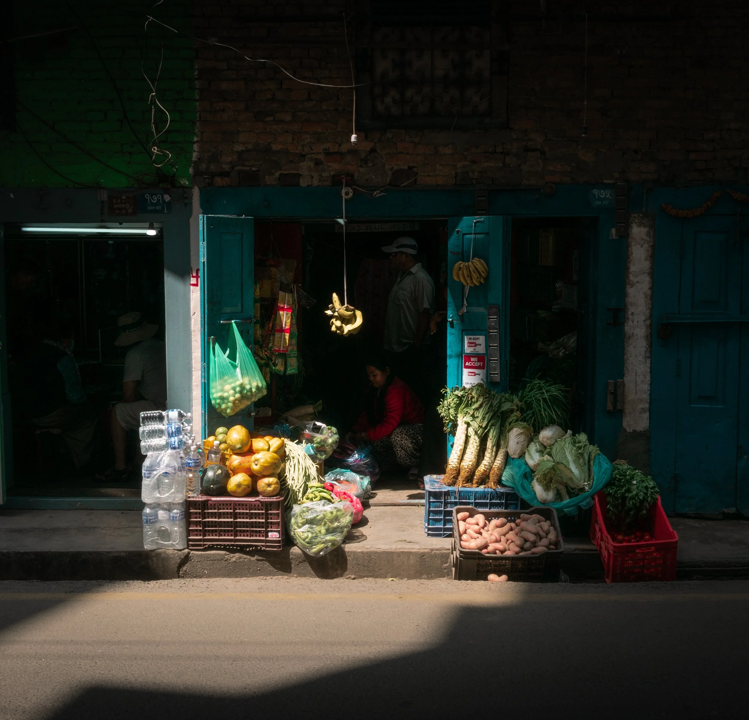 A small outdoor market stall selling fresh fruits and vegetables including bananas, papayas, potatoes, and leafy greens, with water bottles and plastic bags for customers.