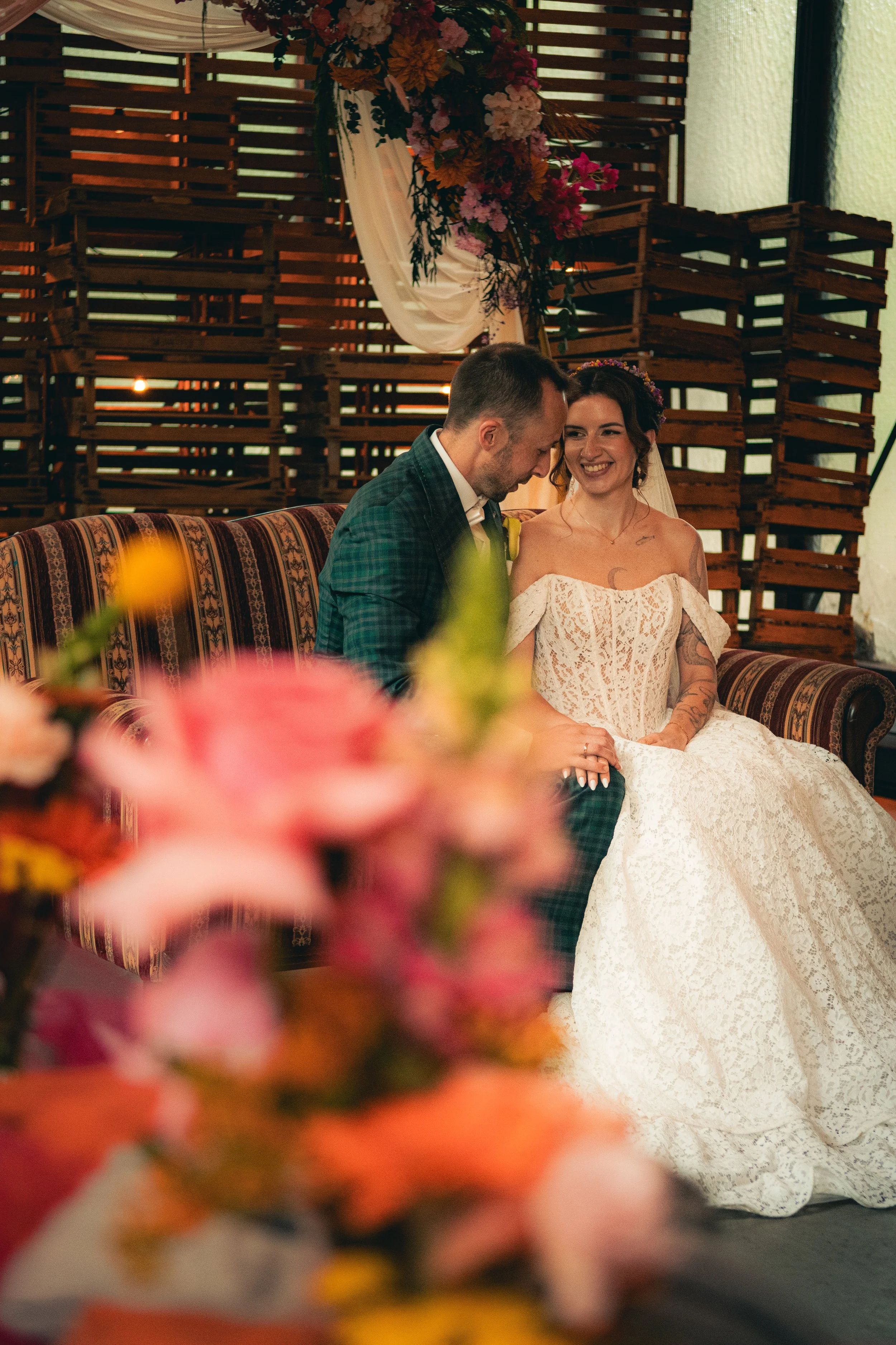 A bride and groom sitting closely together during their wedding ceremony, smiling and enjoying the moment. The bride is wearing a white lace off-shoulder wedding dress and a floral crown, while the groom is dressed in a green plaid suit. The backgrou