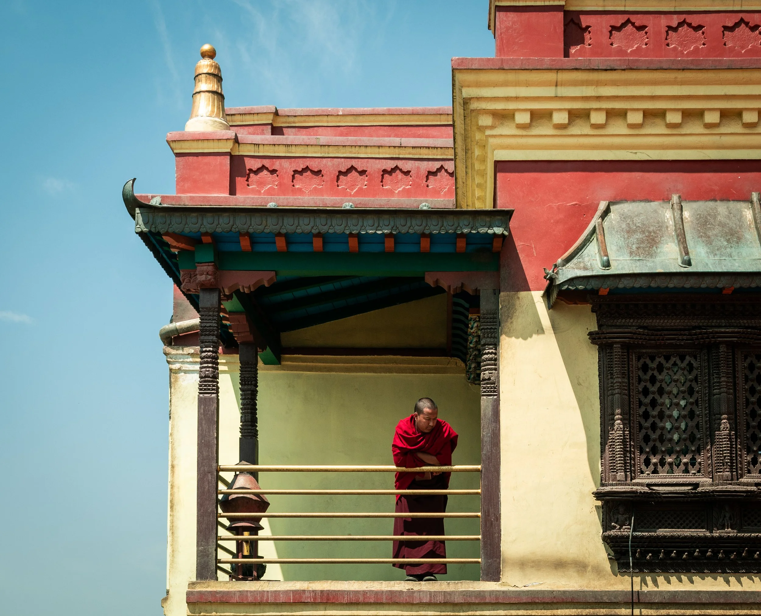 A young monk dressed in red robes standing on the balcony of a traditional building with ornate wooden carvings and a golden finial against a clear blue sky.