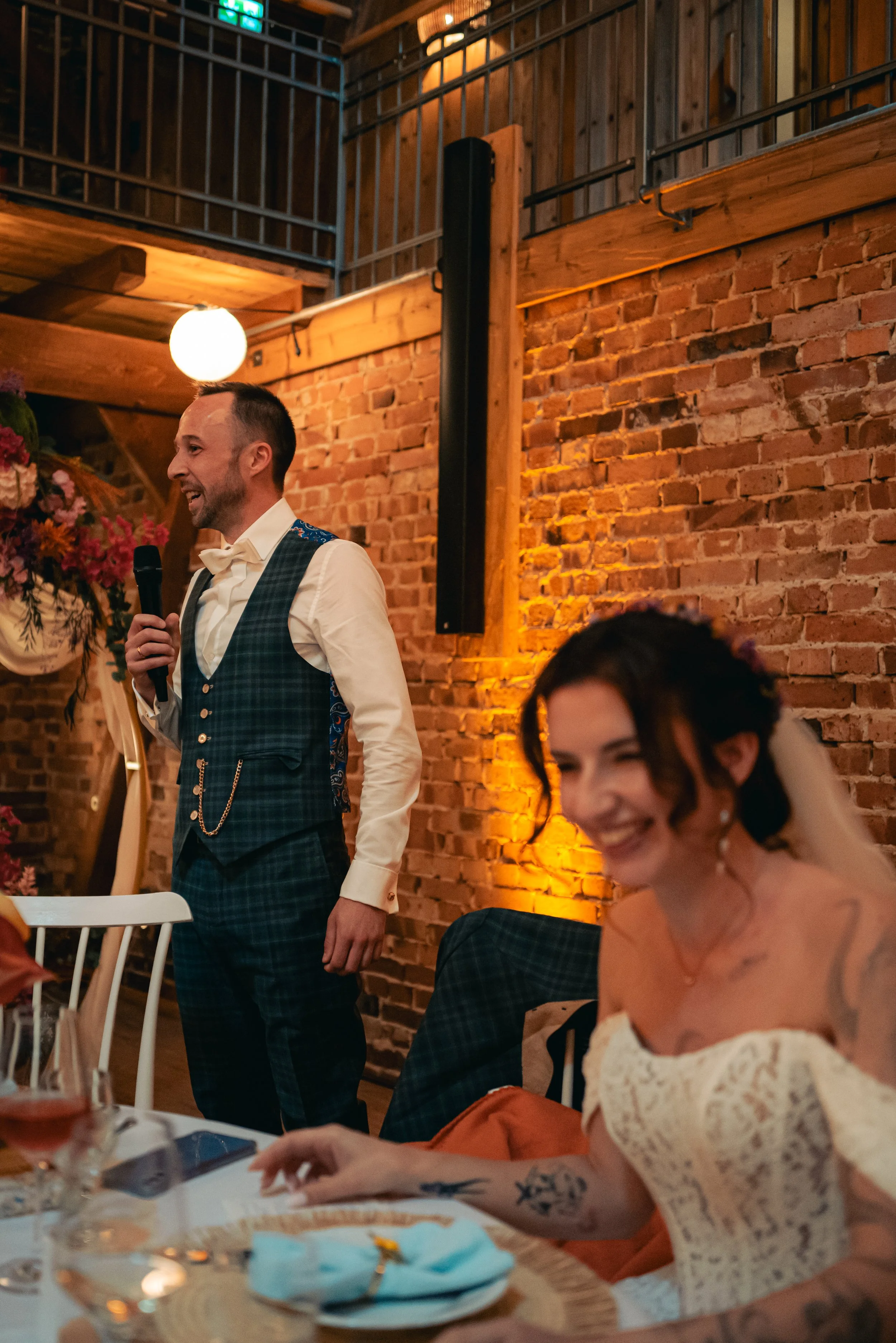 A man in a checkered vest and bow tie speaking into a microphone at a gathering in a rustic venue with exposed brick walls and warm lighting, while a woman with glasses and tattoos smiles at a table in the foreground.