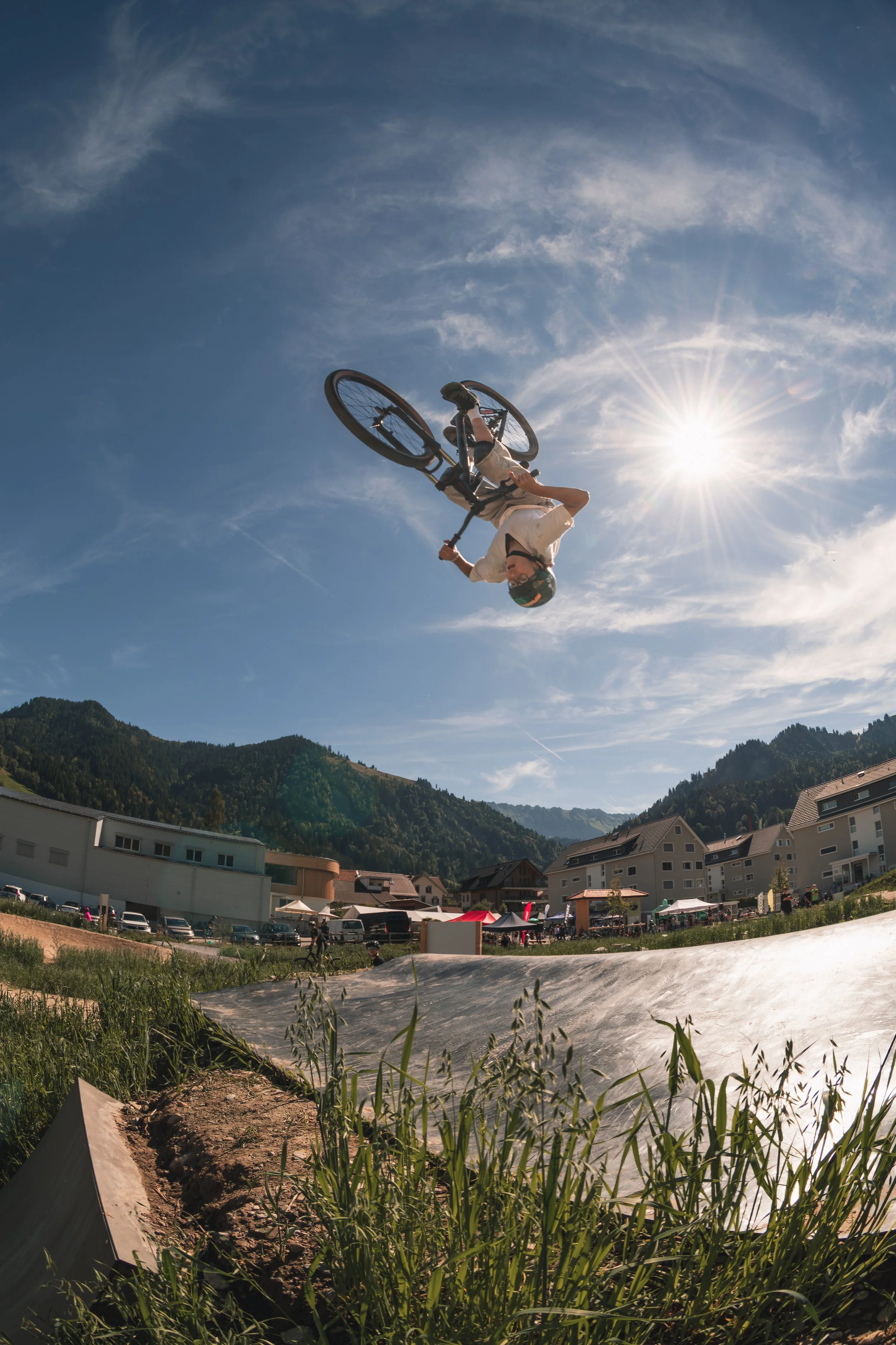 A person performing a backflip on a BMX bike in mid-air at an outdoor biking park with houses and mountains in the background on a sunny day.