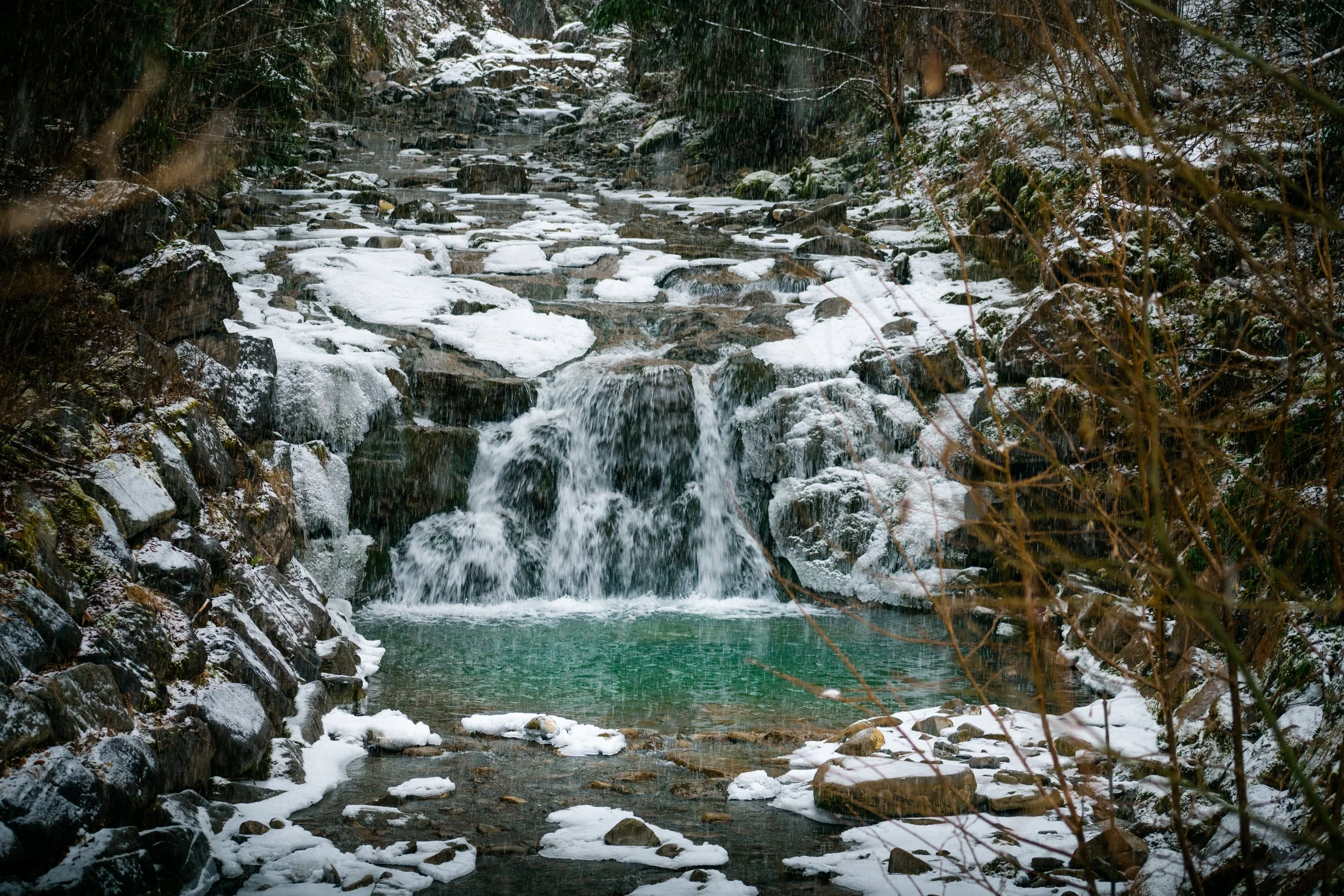 A small waterfall flowing into a clear green pond, surrounded by rocks and snow, with snow-covered trees and bushes in the background.