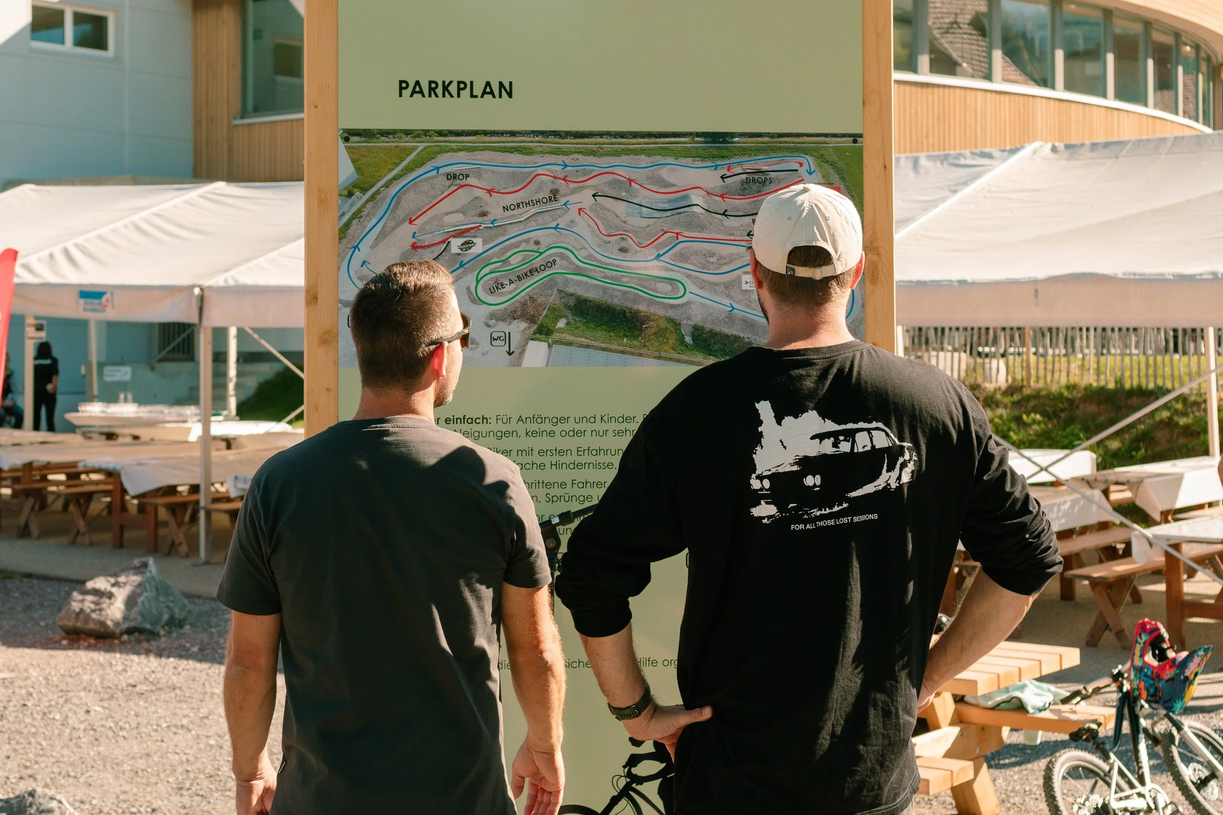 Two men stand in front of a park map, looking at a layout of trails and obstacles, with outdoor seating and a building in the background.