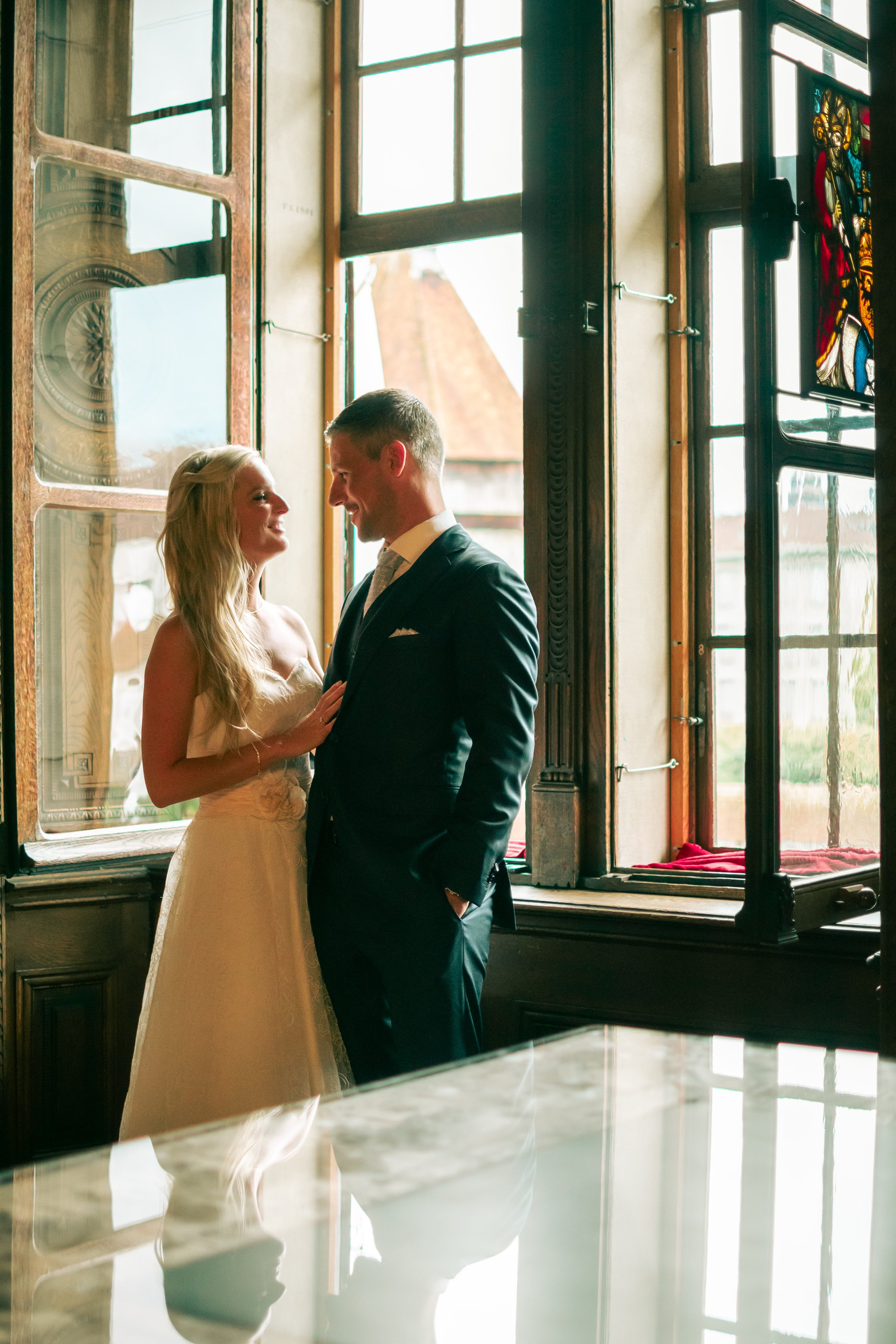A bride and groom standing close together inside near large open windows. The bride is smiling at the groom, and the groom is looking at her, both dressed in wedding attire. The natural light from outside illuminates the scene, with a church steeple 