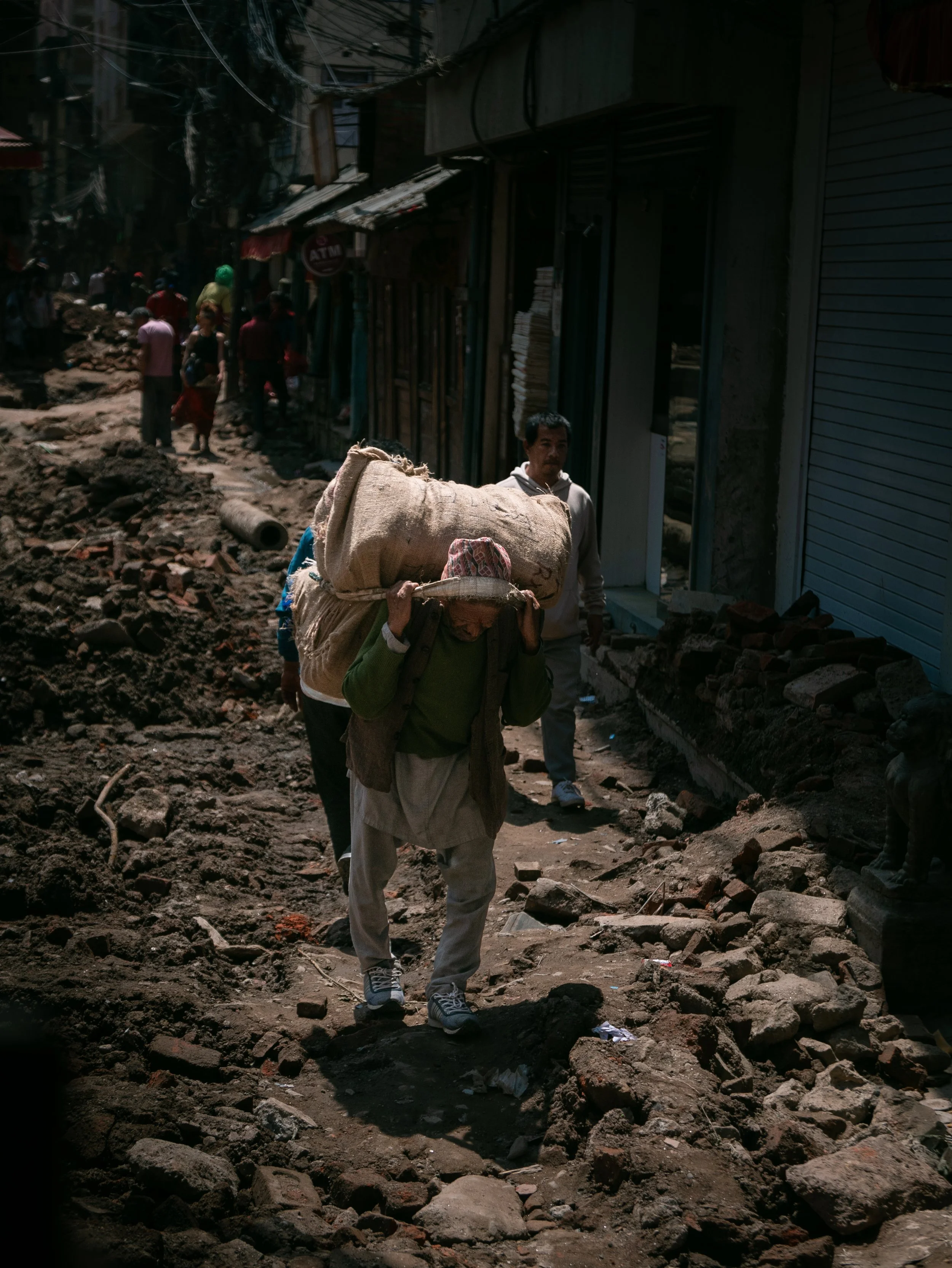 People walking on a muddy, debris-strewn street in an urban area with damaged buildings and overhead wires.