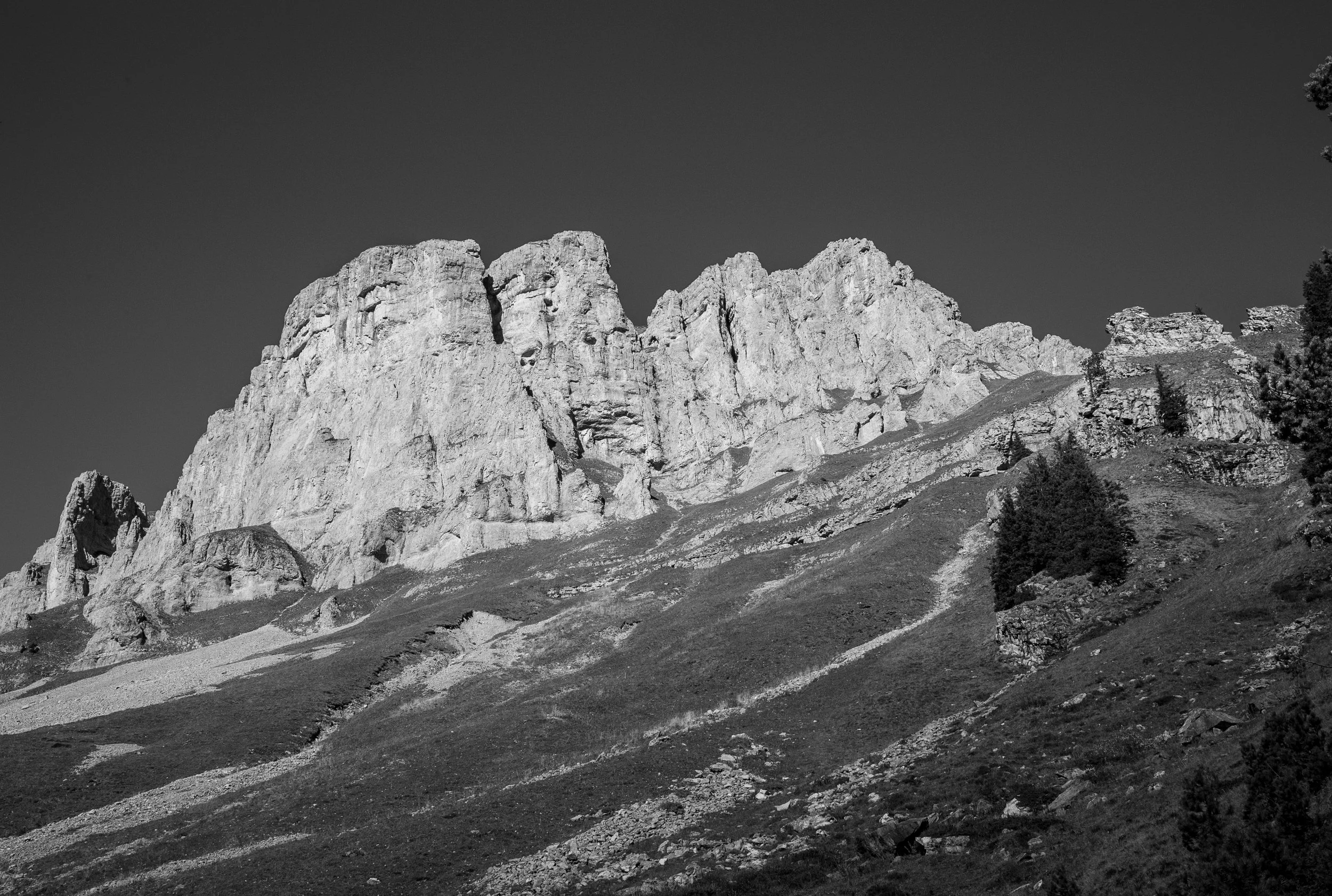 Black and white photograph of rugged mountain peaks with sparse trees at the base, under a clear sky.