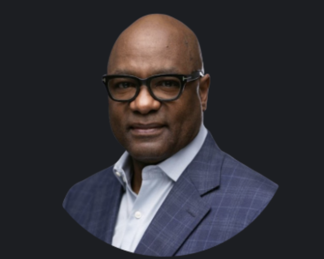 A portrait of an African American man wearing black glasses, a white shirt, and a dark blue blazer with a checkered pattern, against a dark background.