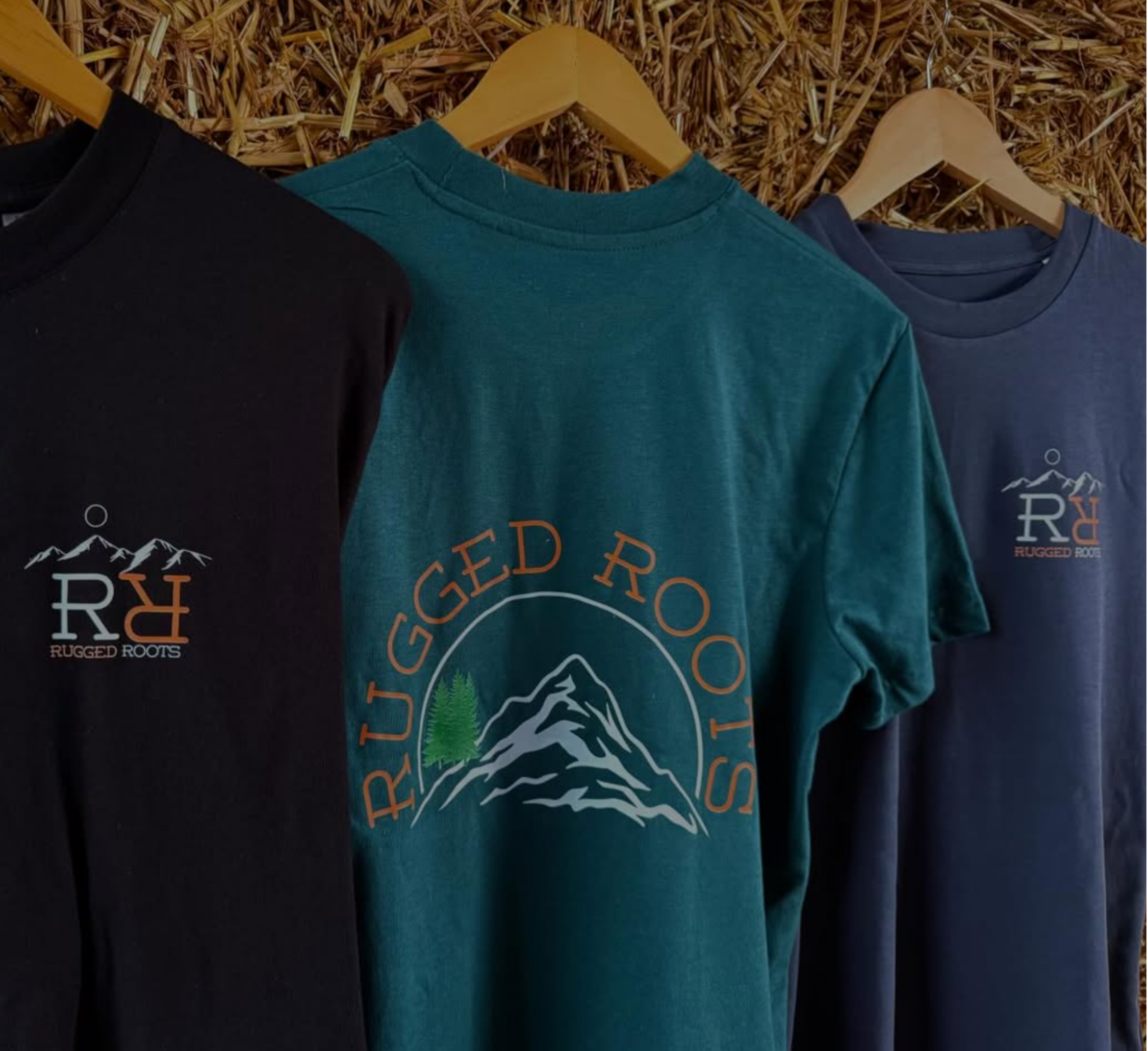 Three t-shirts are hanging on wooden hangers, displayed against a background of dried grass or straw. The t-shirts are in black, teal, and blue colors, each with a logo that features the words "Rugged Roots" and an illustration of mountain peaks and trees.