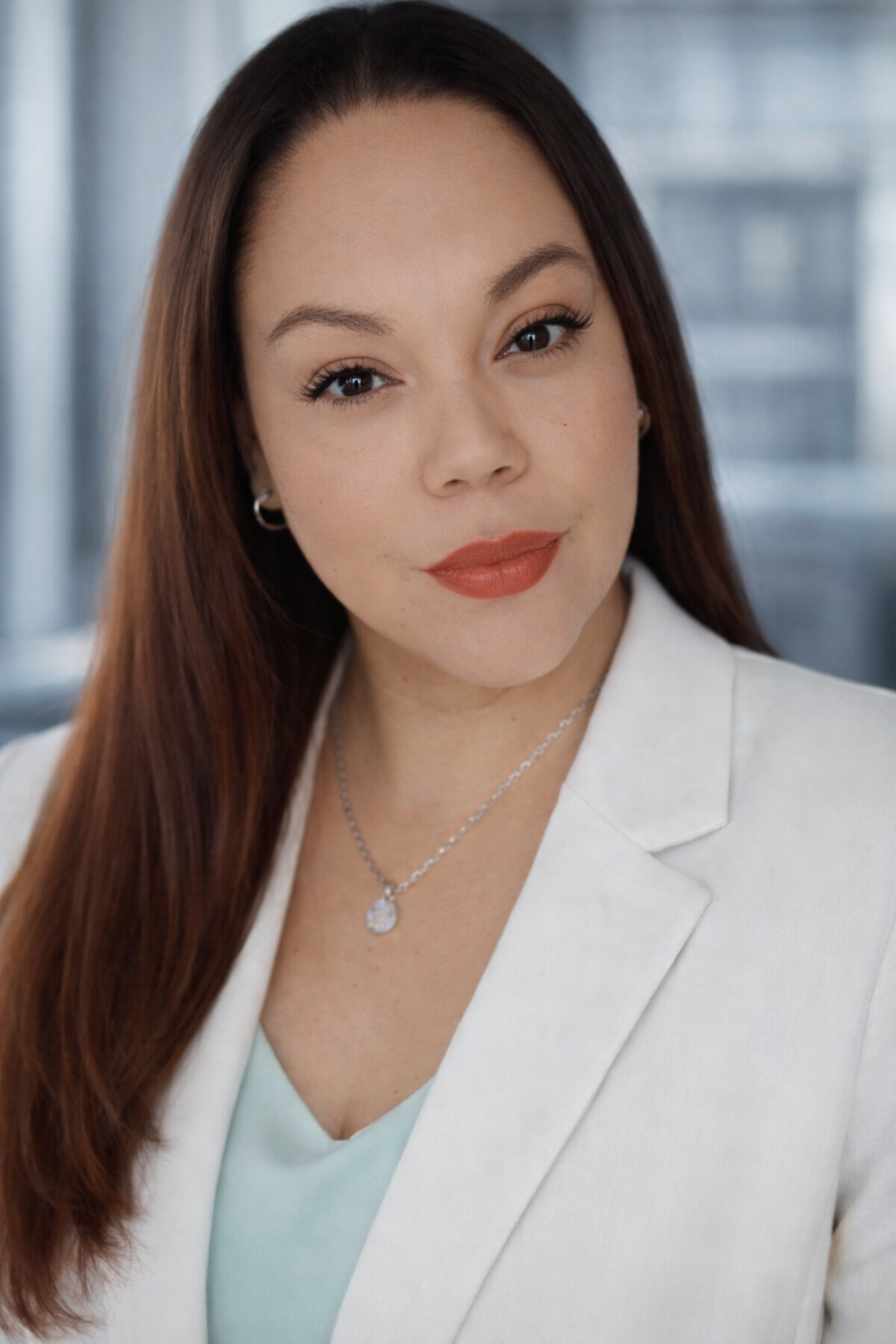 Professional woman with long brown hair and red lipstick, wearing a white blazer and a necklace. She is looking at the camera with a slight smile. Blurred office background.