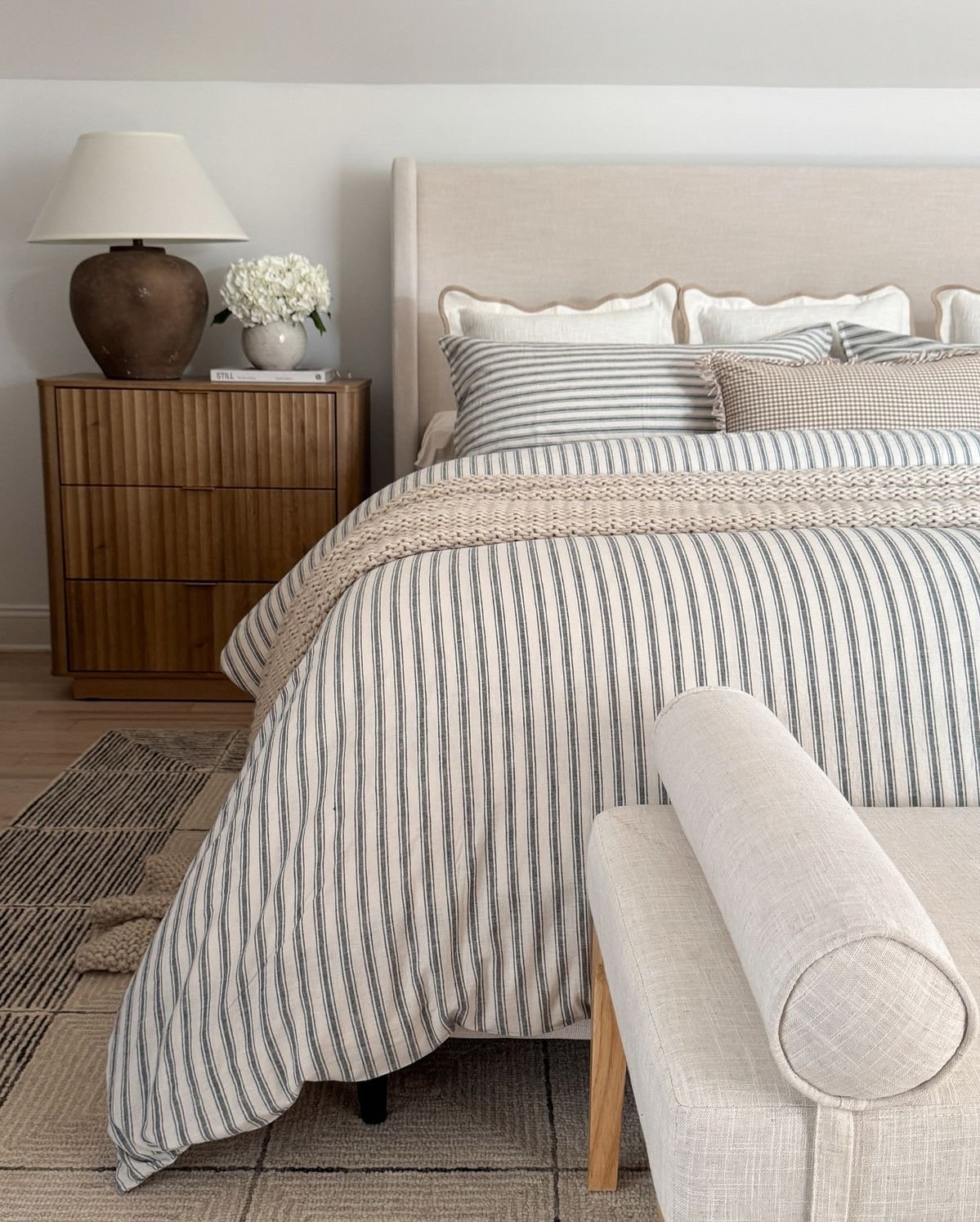 Modern coastal bedroom design featuring a beige linen winged headboard, blue and white striped bedding, a textured knit throw, and an organic fluted wood nightstand with a concrete base lamp.