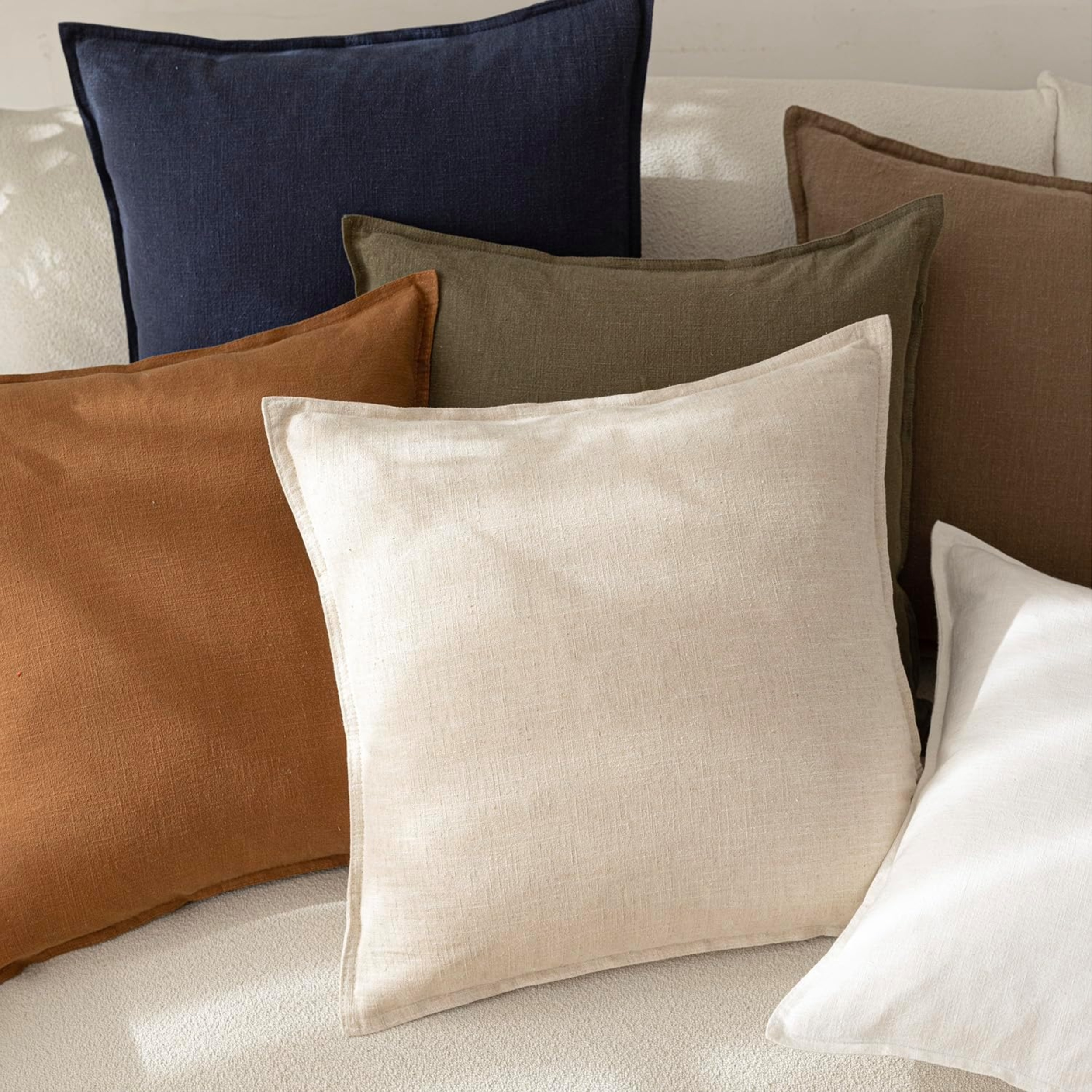 A curated stack of linen throw pillows in a modern organic color palette, featuring shades of terracotta, olive green, navy blue, charcoal, and creamy oatmeal on a white textured bouclé sofa.