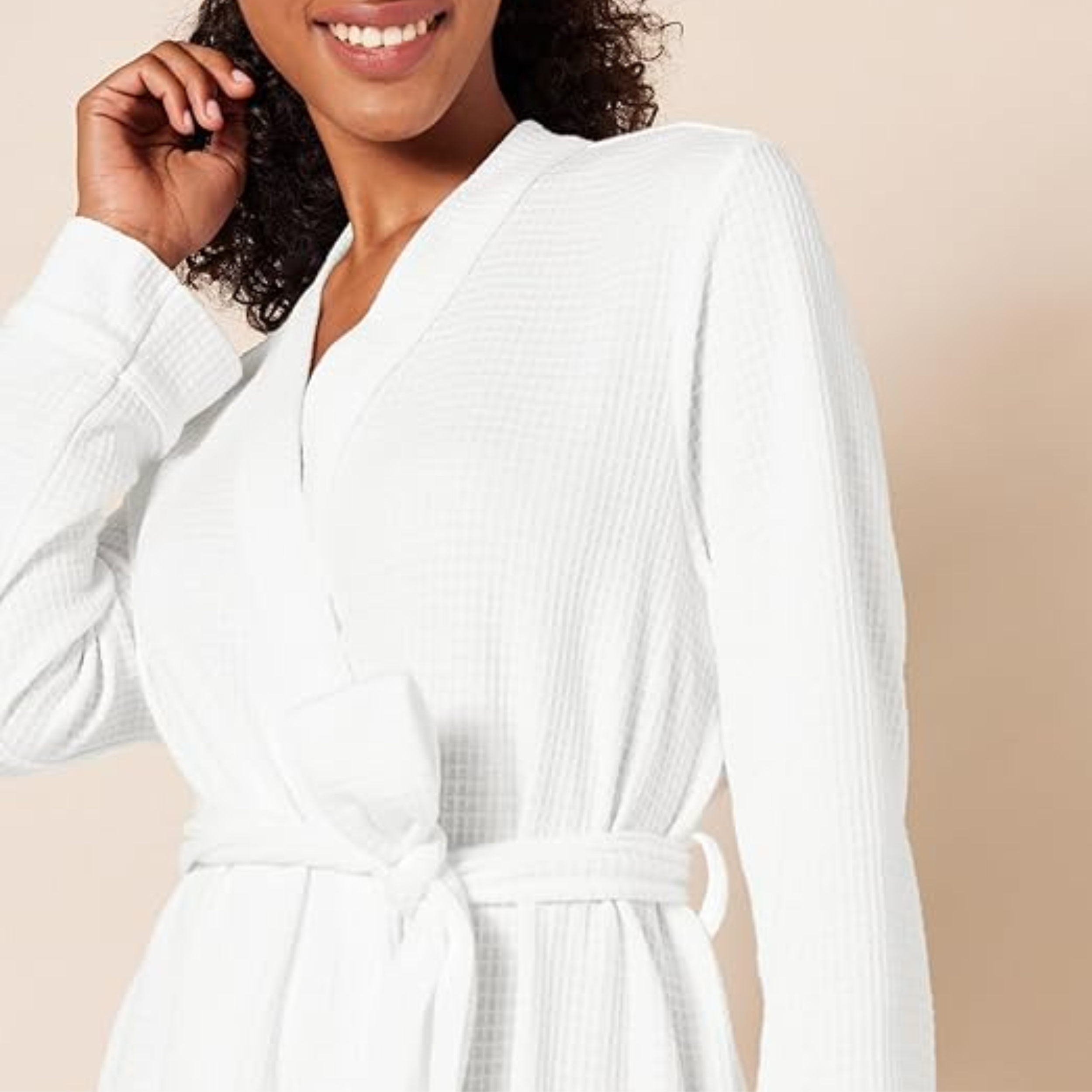 A smiling woman with dark curly hair from the shoulders down, wearing a belted, plush white waffle knit bathrobe, with one hand near her neck.
