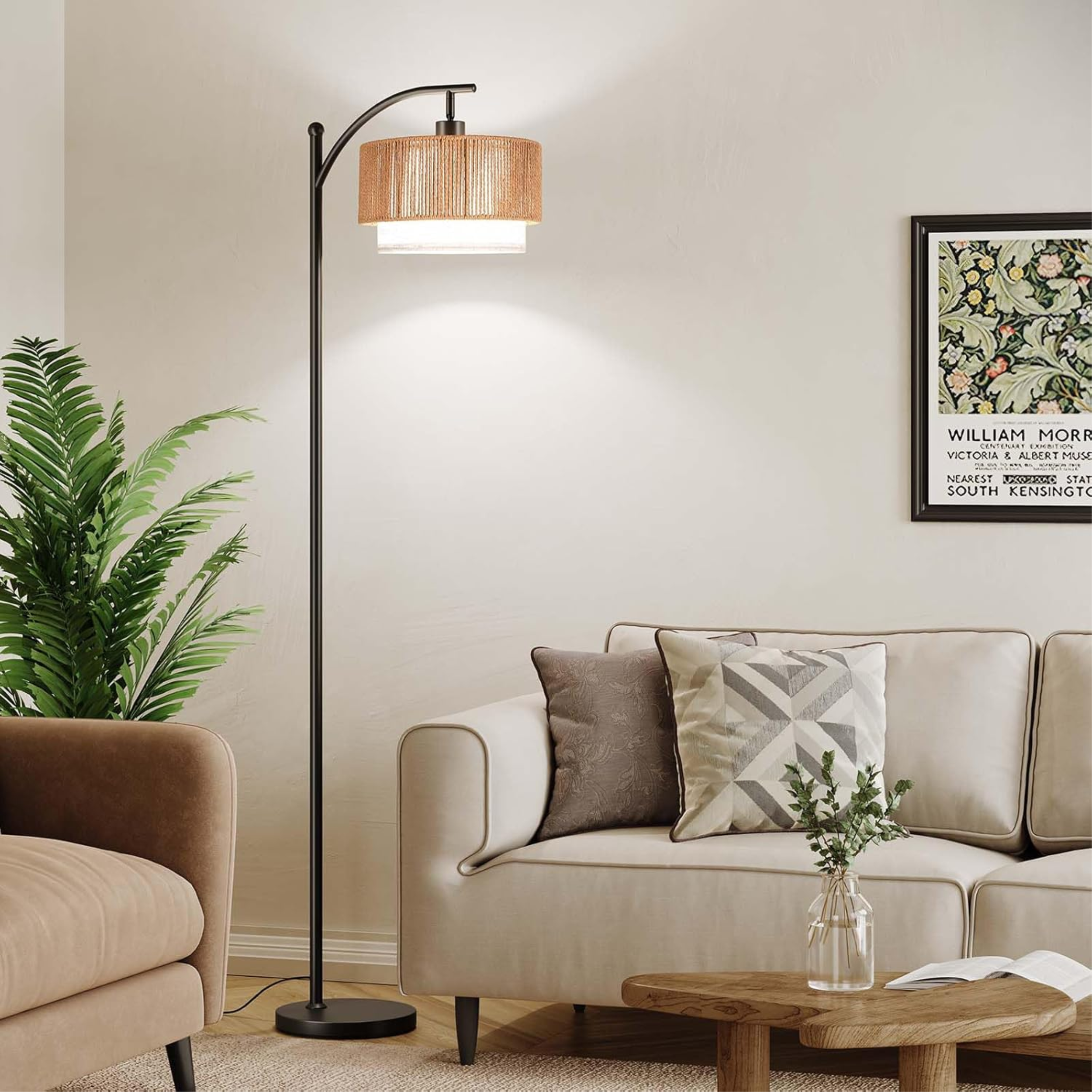 A modern black arc floor lamp with a dual-layered rattan and fabric drum shade positioned over a cream sofa. The room features a William Morris art print, a lush green floor plant, and an oak coffee table, embodying an organic modern aesthetic.