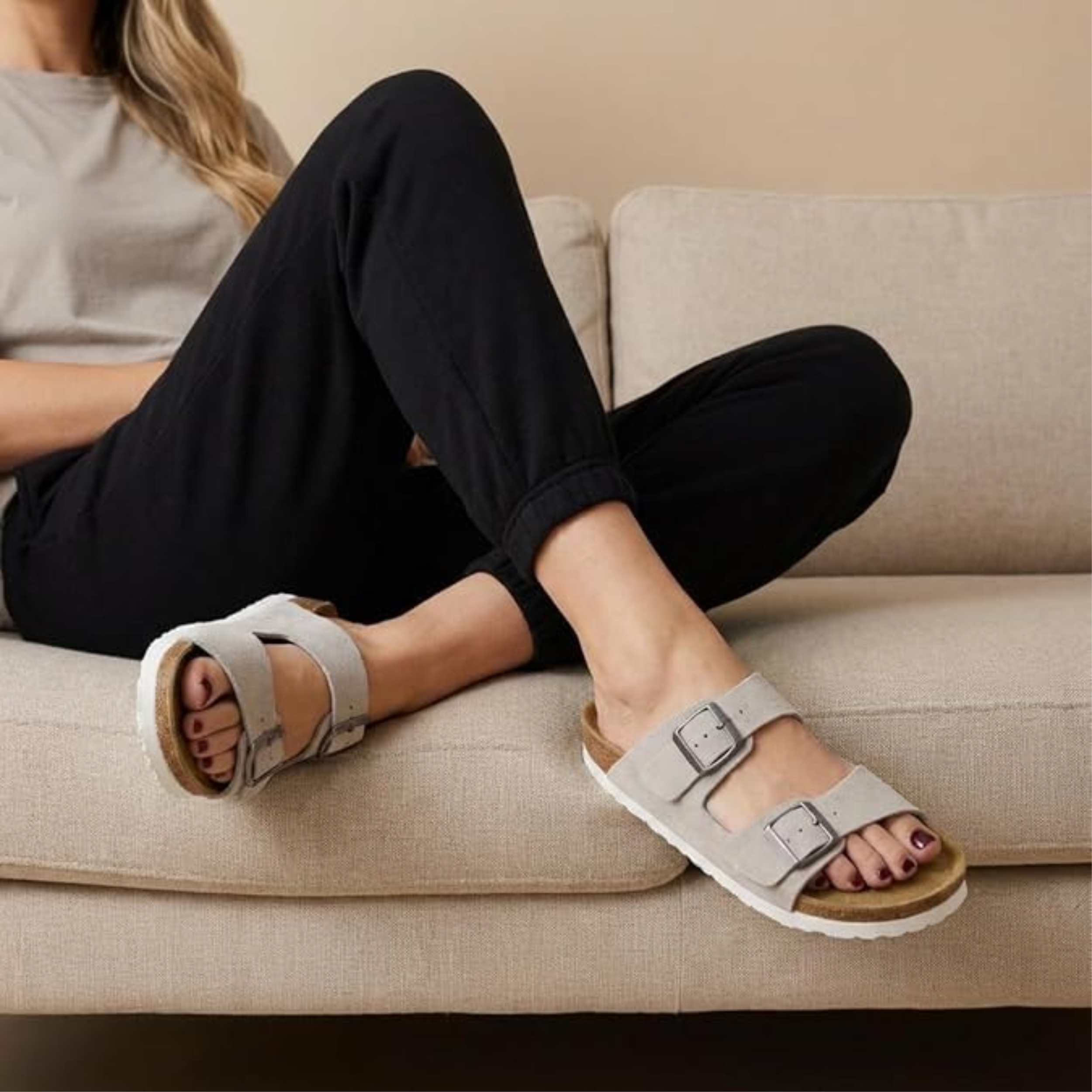 Close-up of a person's feet and lower legs while sitting on a light-colored sofa, showcasing beige suede two-strap sandals with a contoured cork footbed and white sole.
